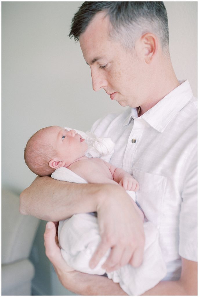 Father holds his newborn baby swaddled in white up close to him as the baby looks back at him.