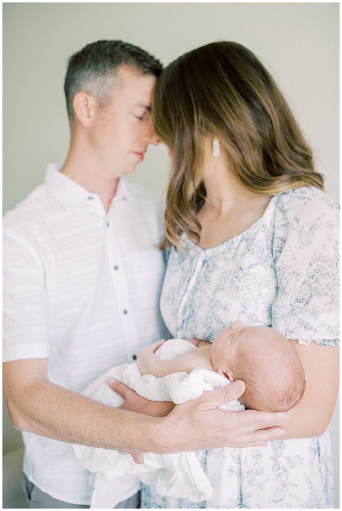 Mother and father lean their heads into one another and close their eyes while holding their newborn baby.