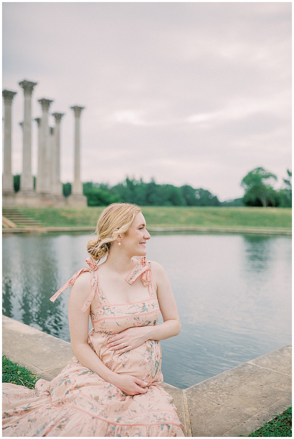 Blonde mother in Zimmerman pink dress sits in front of pond with hands on her belly during her maternity session.