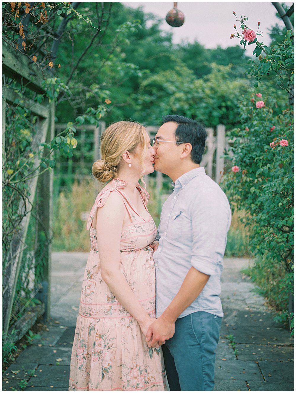 Husband and wife face each other and kiss during their National Arboretum maternity session under floral arch.
