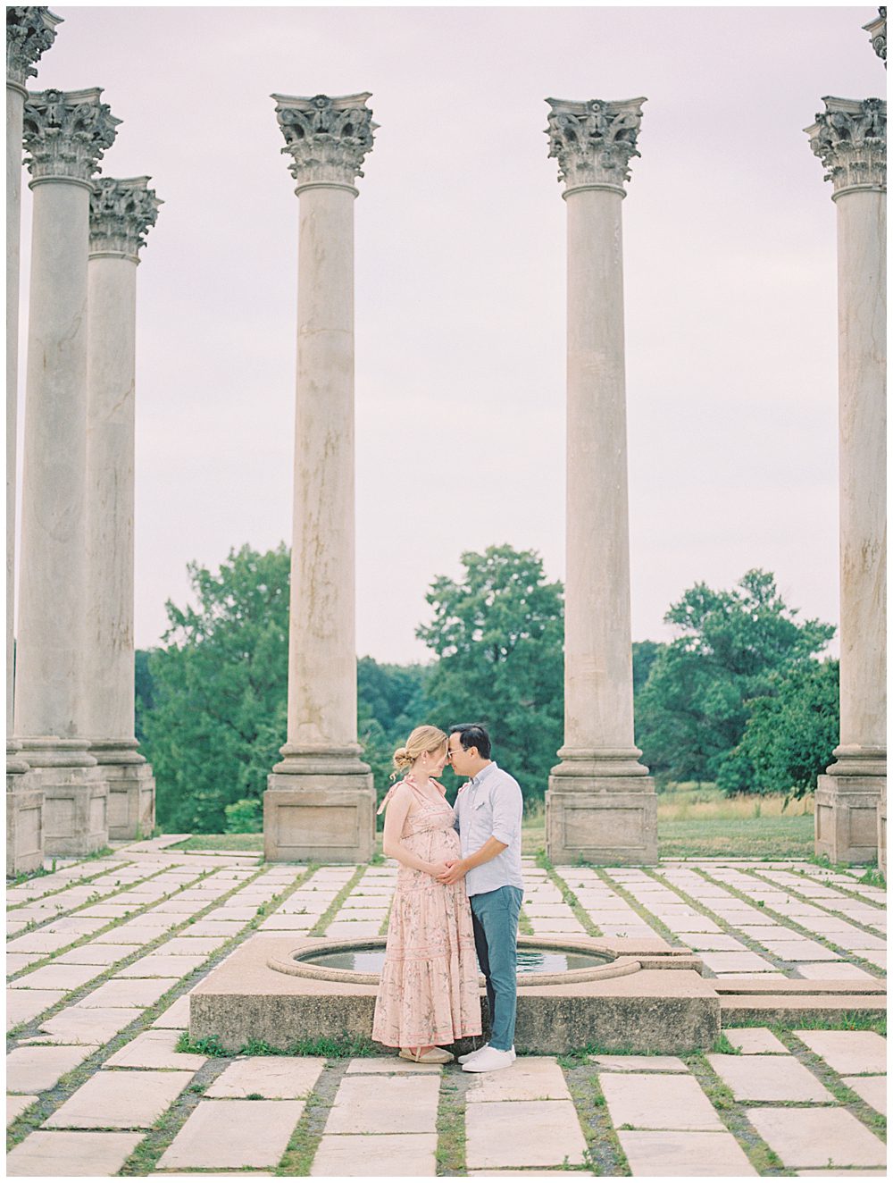 Expecting parents stand in the center of the National Arboretum columns, facing one another during their maternity session.