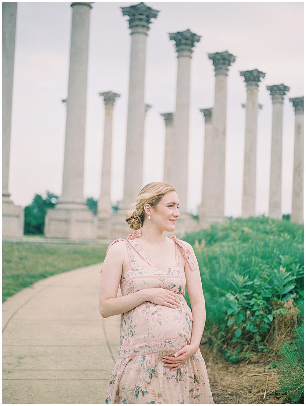 Pregnant mother in pink Zimmerman dress stands in front of National Arboretum columns with hands on her belly as the wind blows.