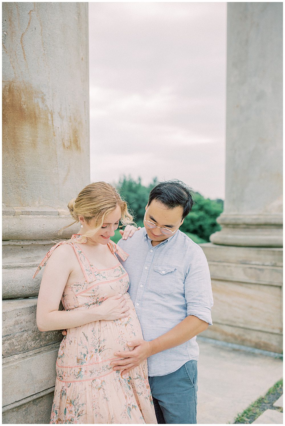 Pregnant mother leans against pillar while husband places hand on her belly during their National Arboretum maternity session.