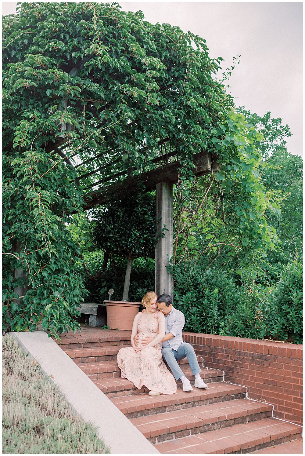 Husband and wife sit on brick steps underneath pergola at the National Arboretum during maternity session.