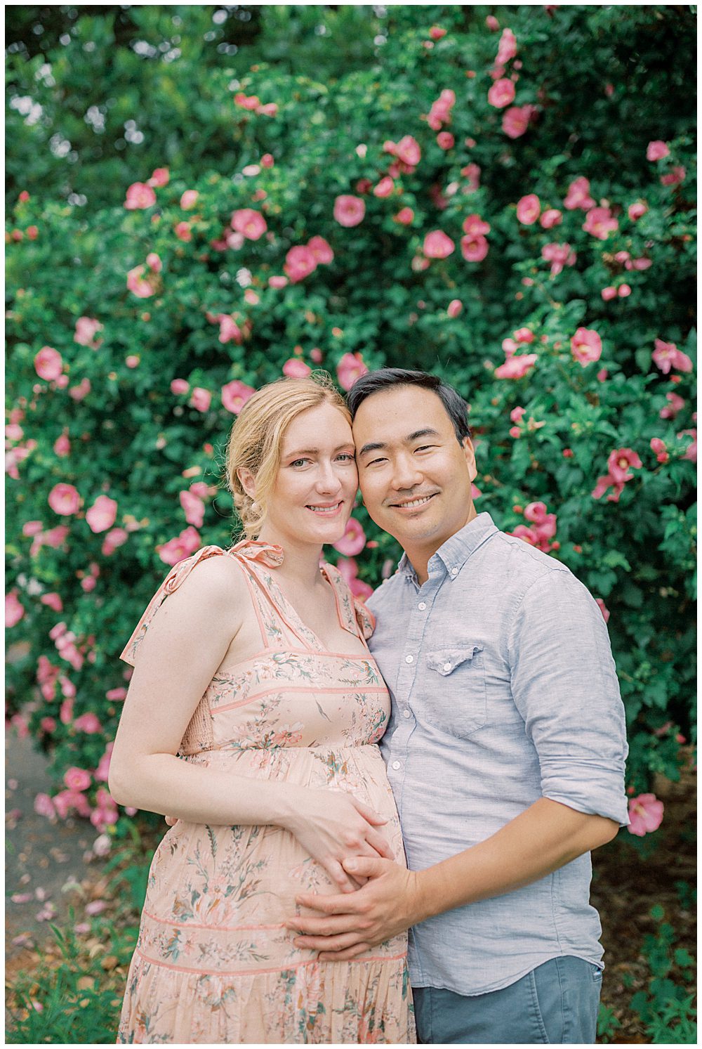 Expecting parents lean into one another and smile while standing in front of a rose bush.