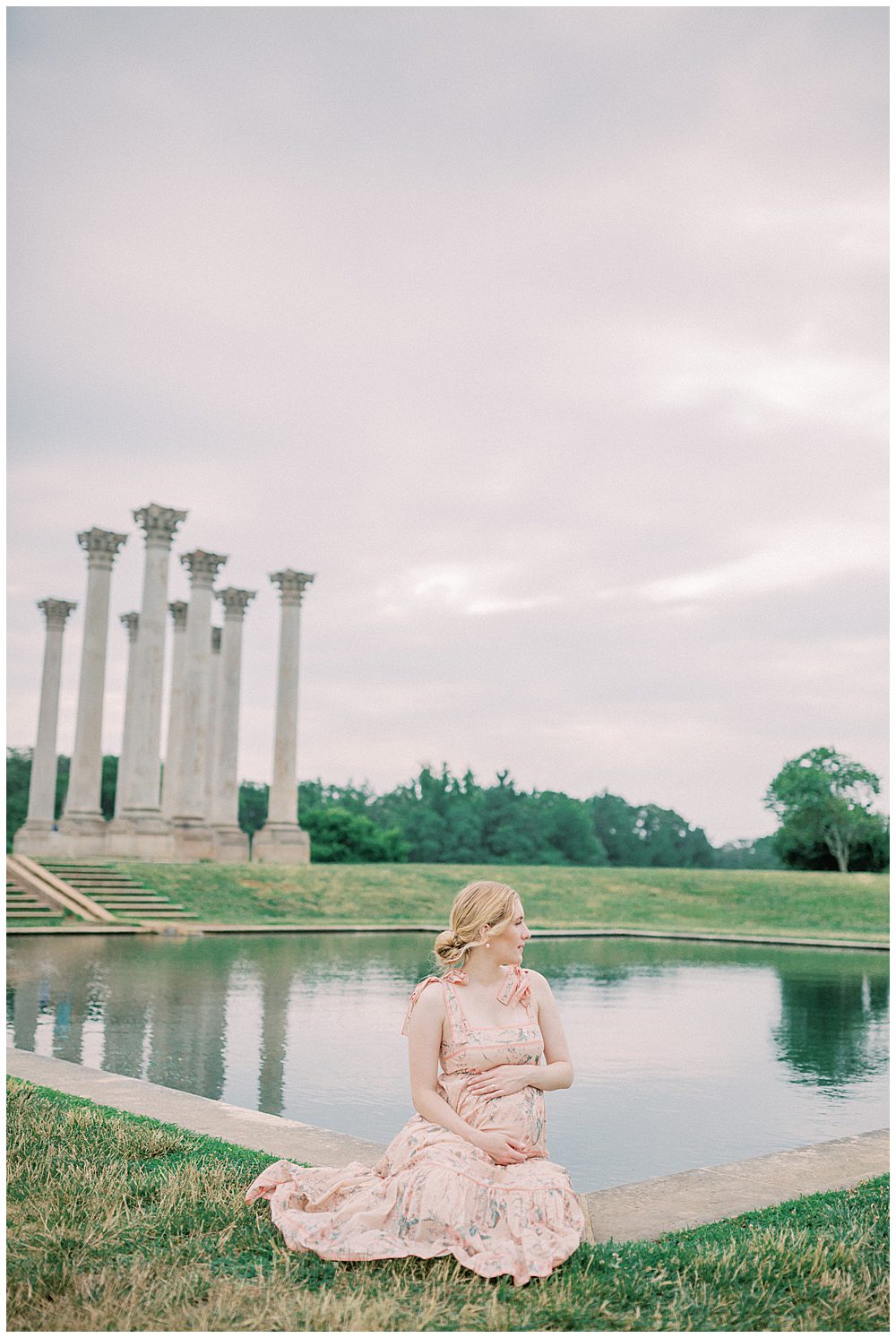 Pregnant woman sits in front of National Arboretum pond with hands on belly, looking out into the distance.