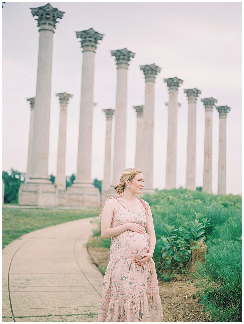 Pregnant blonde mother stands in front of National Arboretum columns with hands on belly during maternity session as her hair blows in wind.