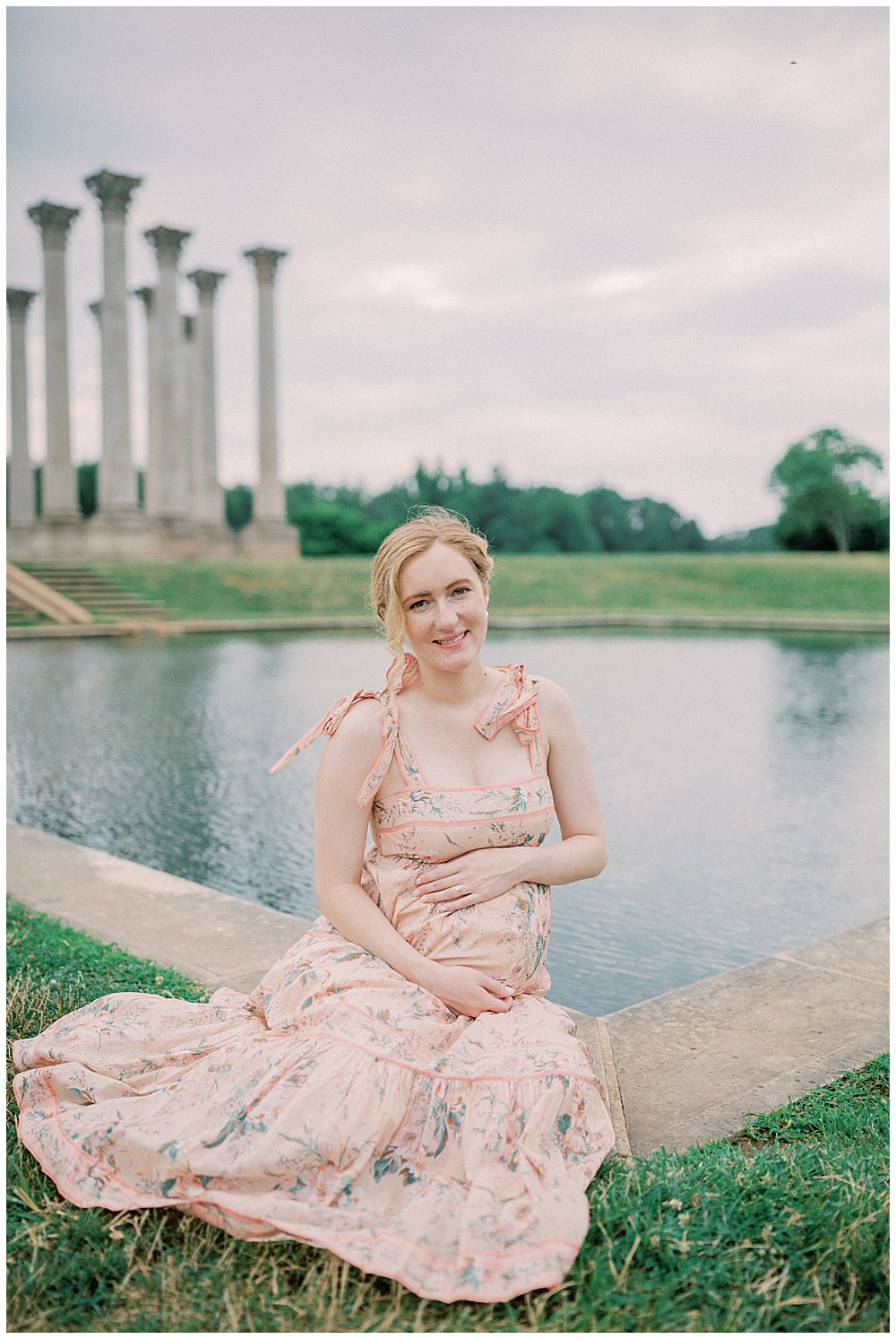 Pregnant blonde mother sits in front of pond at the National Arboretum and smiles at the camera.