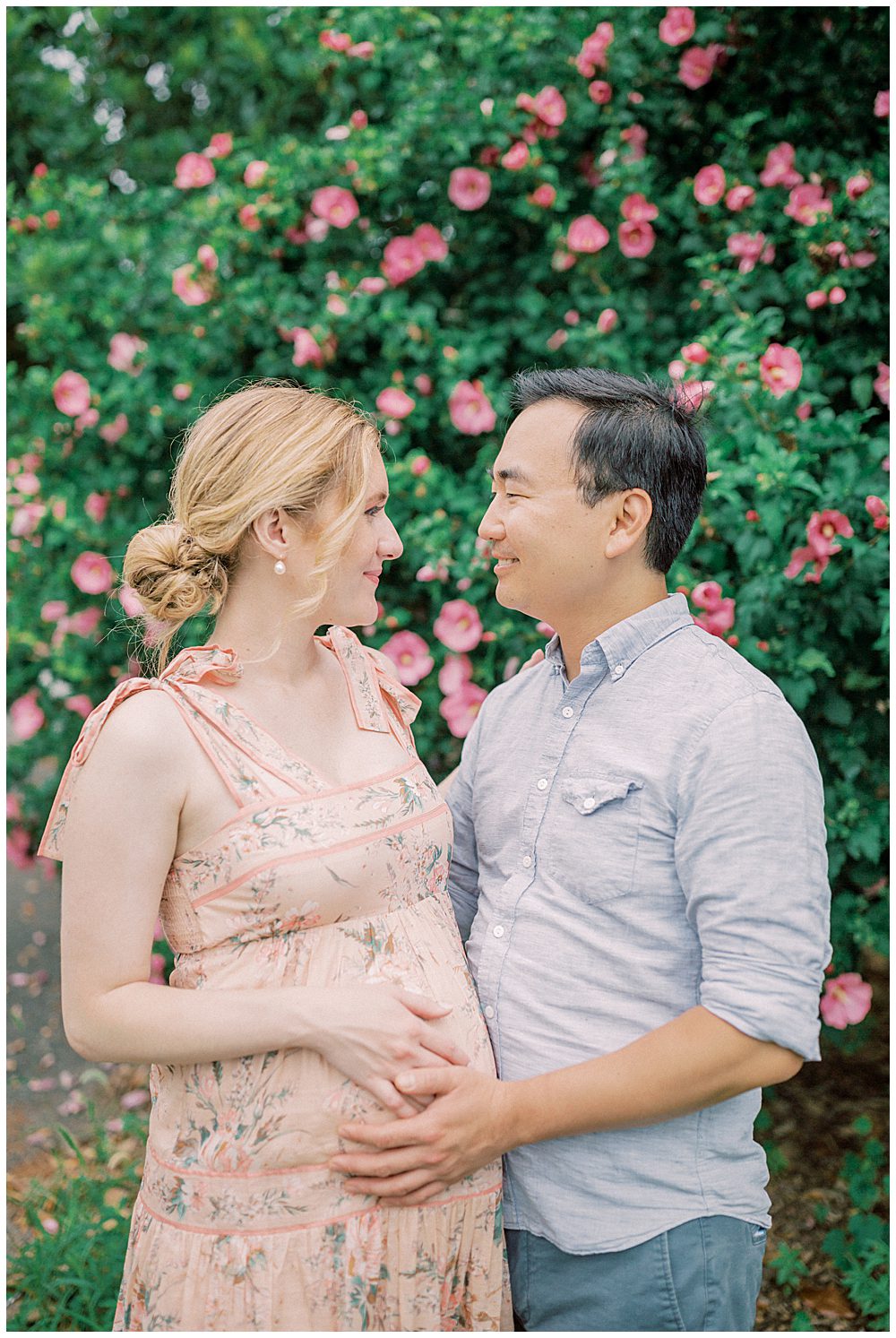 Pregnant woman in pink Zimmerman dress faces her husband as they stand in front of a Rose of Sheron bush during their National Arboretum maternity session.