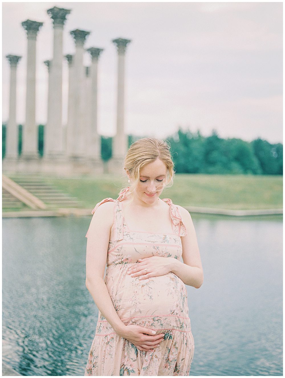 Pregnant mother places one hand on top and one hand below belly while standing in front of National Arboretum pond during National Arboretum maternity session.