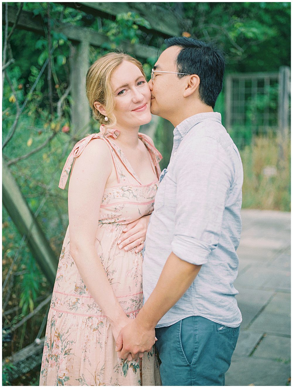 Husband kisses his wife's cheek as she smiles as they stand under a floral arch.