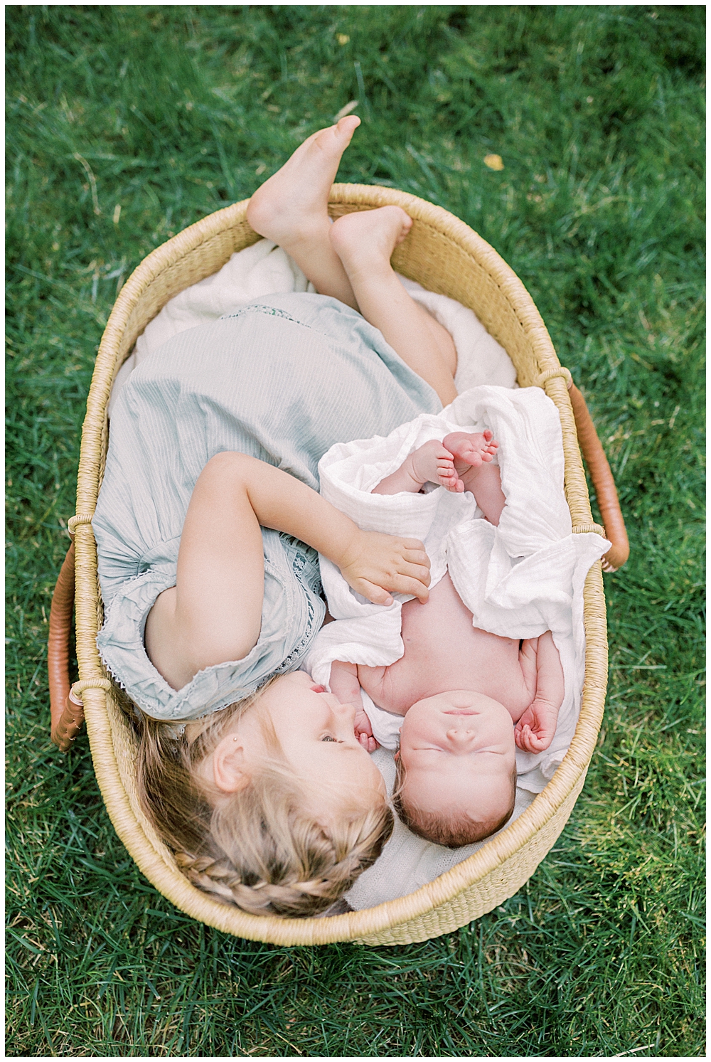 Toddler girl lays in Moses basket with her newborn baby sister during outdoor studio newborn session