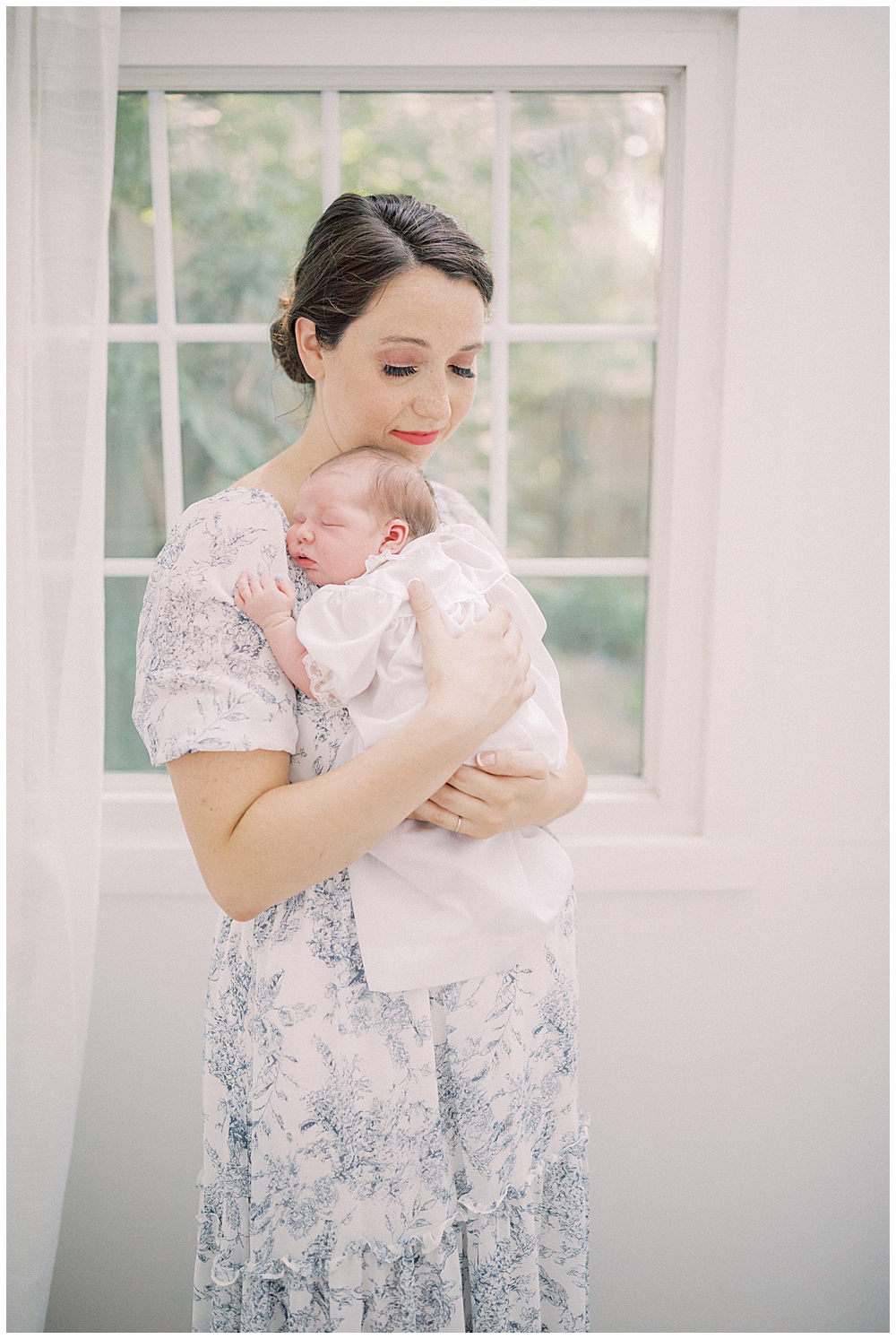 M other holds her newborn daughter up to her chest while standing in front of a window