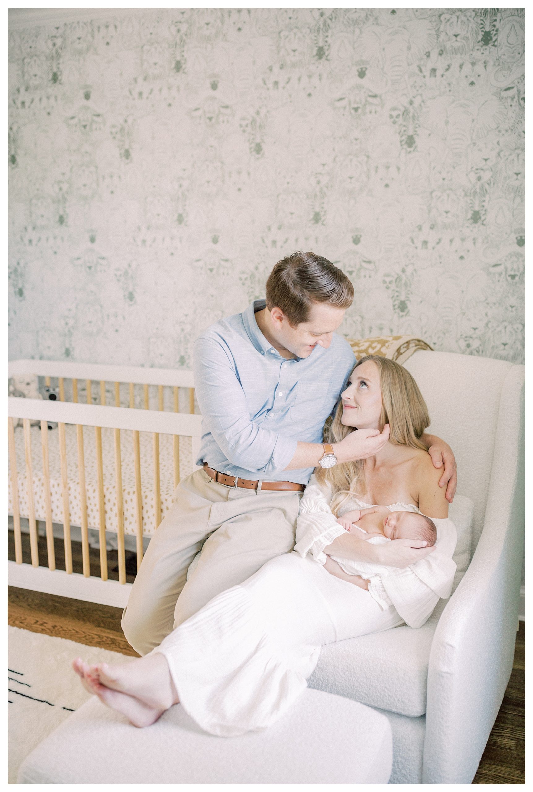 New father sits on edge of chair in nursery, caressing his wife's cheek who holds their newborn.