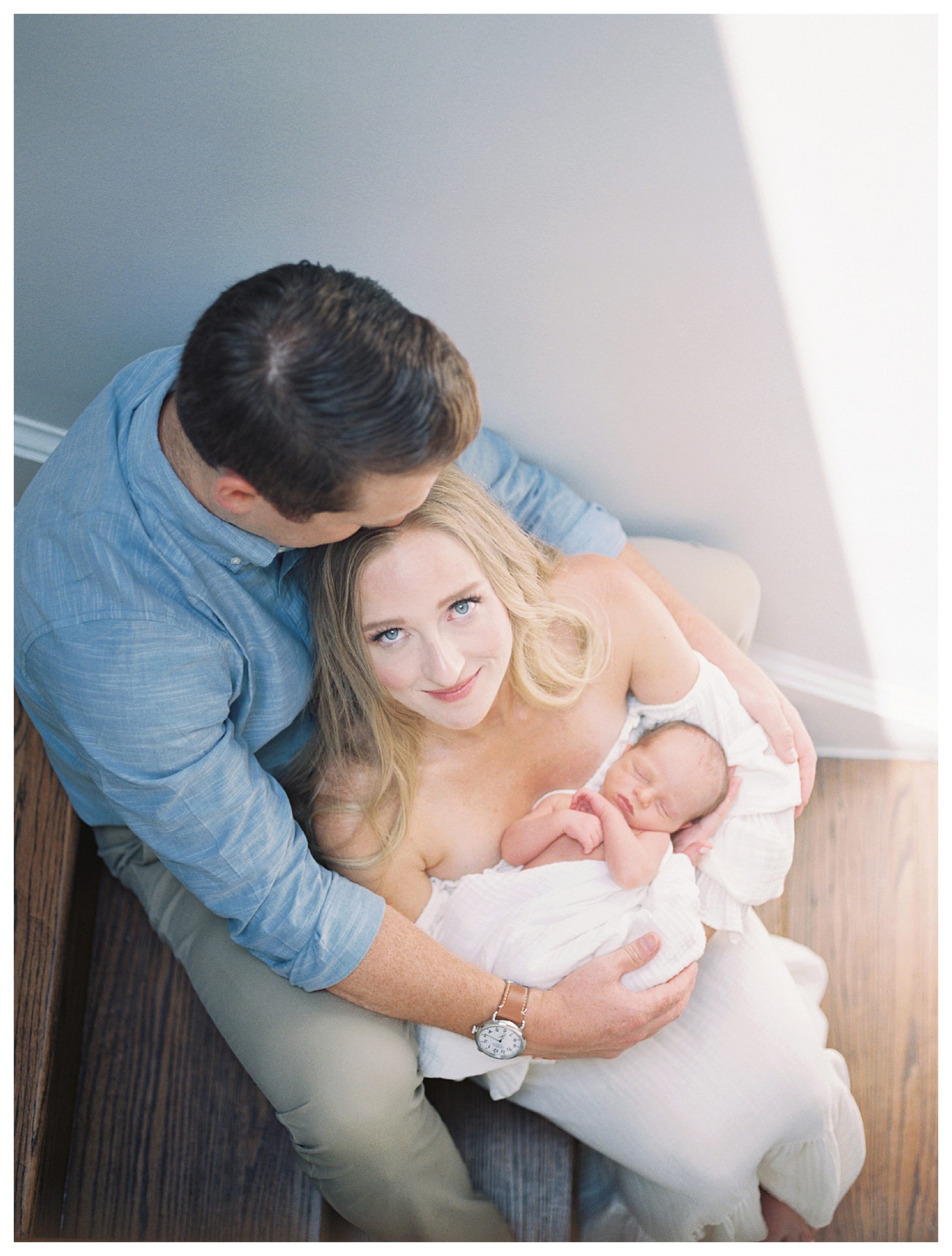 Blonde mother sits on steps with husband holding newborn baby and looks at the camera.