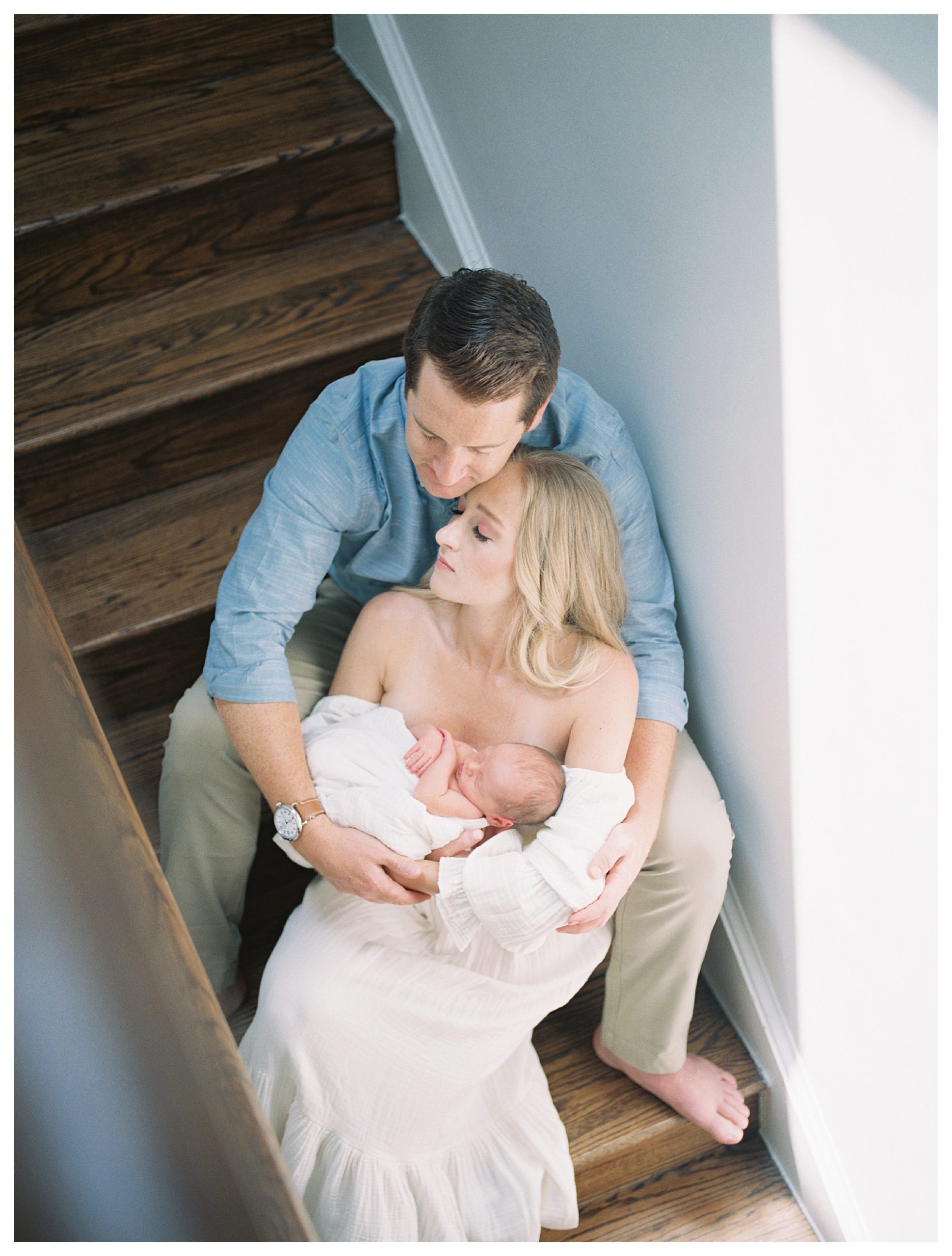 Father and mother sit on steps, leaning into one another as they hold their newborn baby.