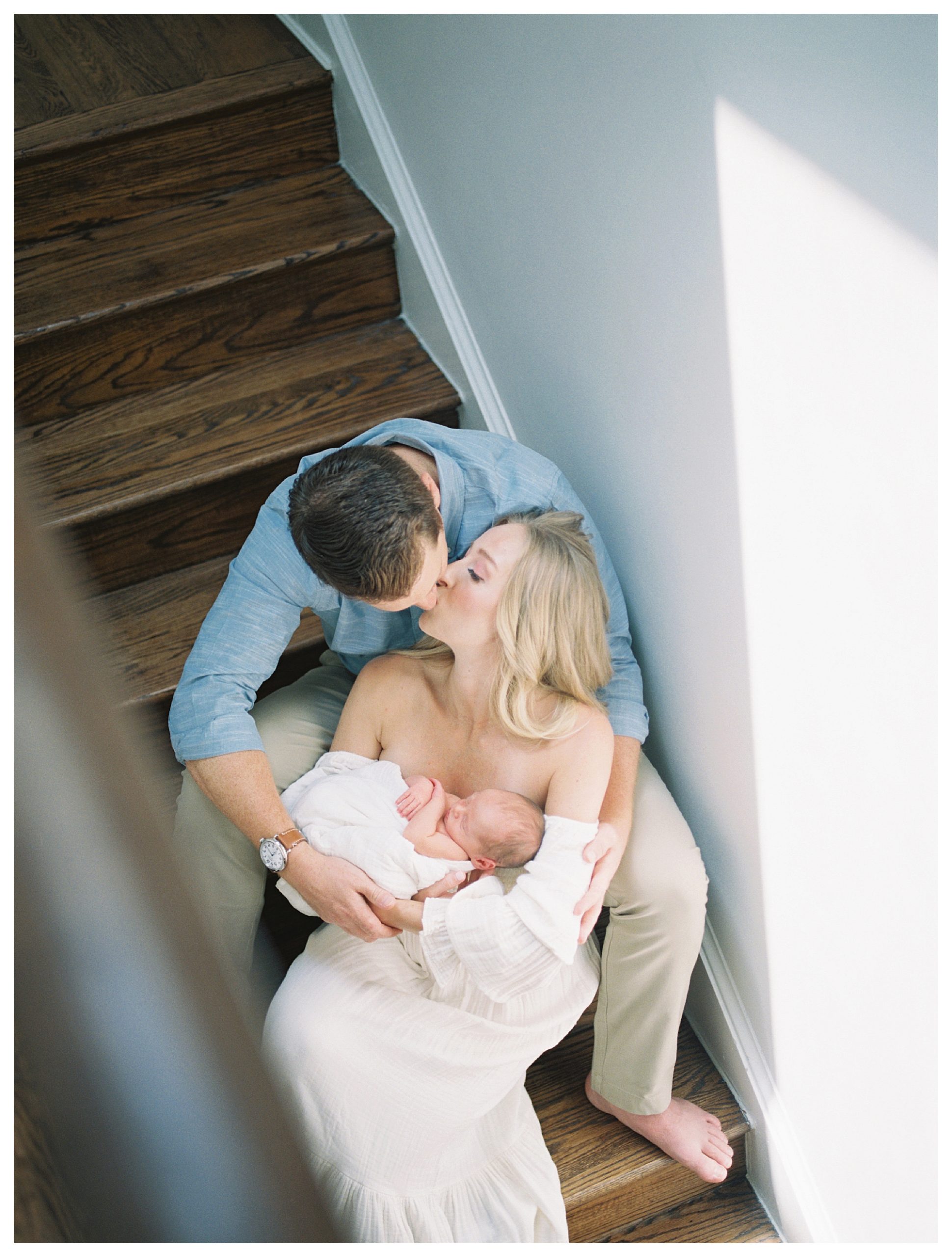 Mother and father sit on steps and kiss while holding newborn baby.