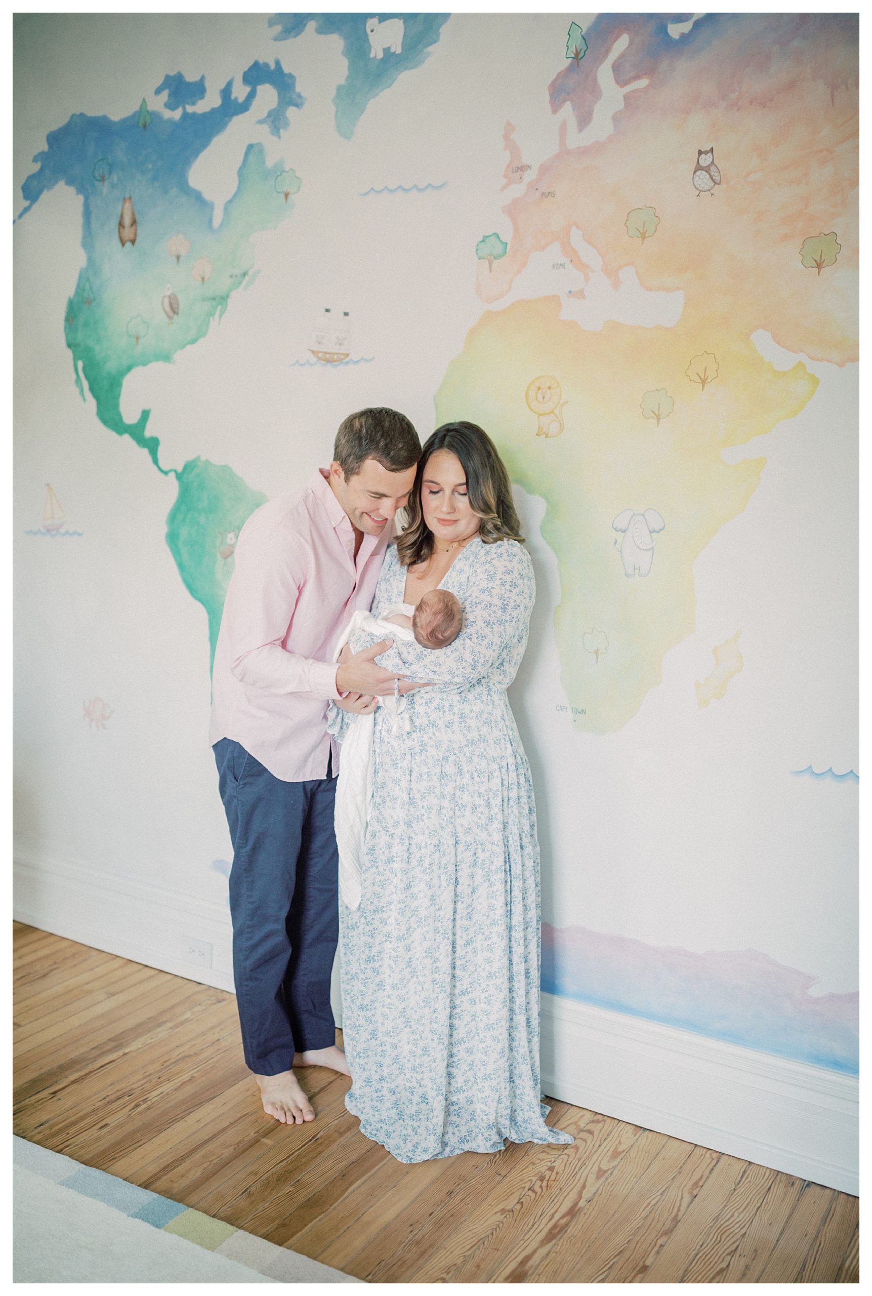 Father leans over and smiles at newborn baby held by mother as they stand in front of colorful painted mural in the nursery.