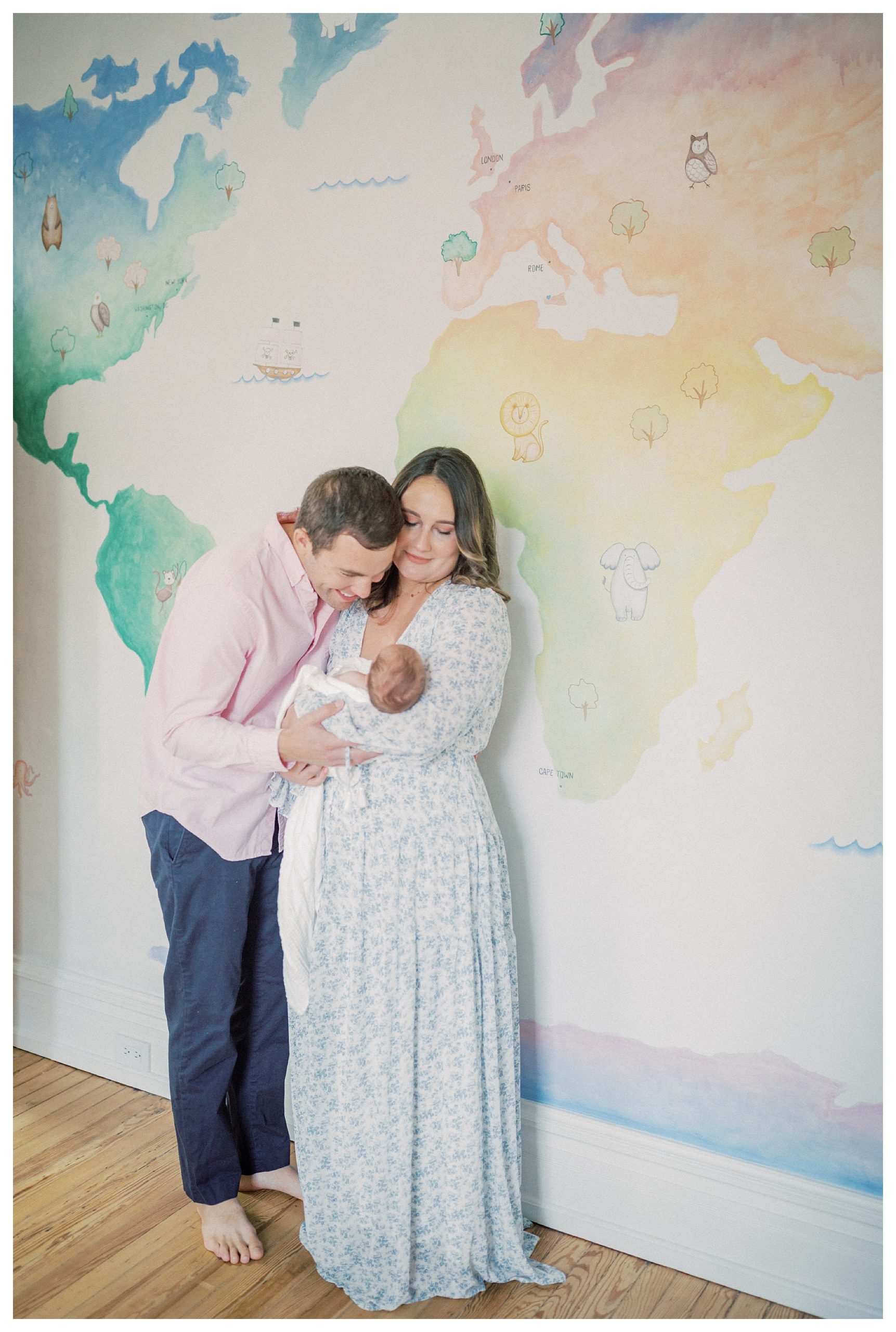 Father leans over and smiles at newborn baby held by mother as they stand in front of painted mural in nursery.