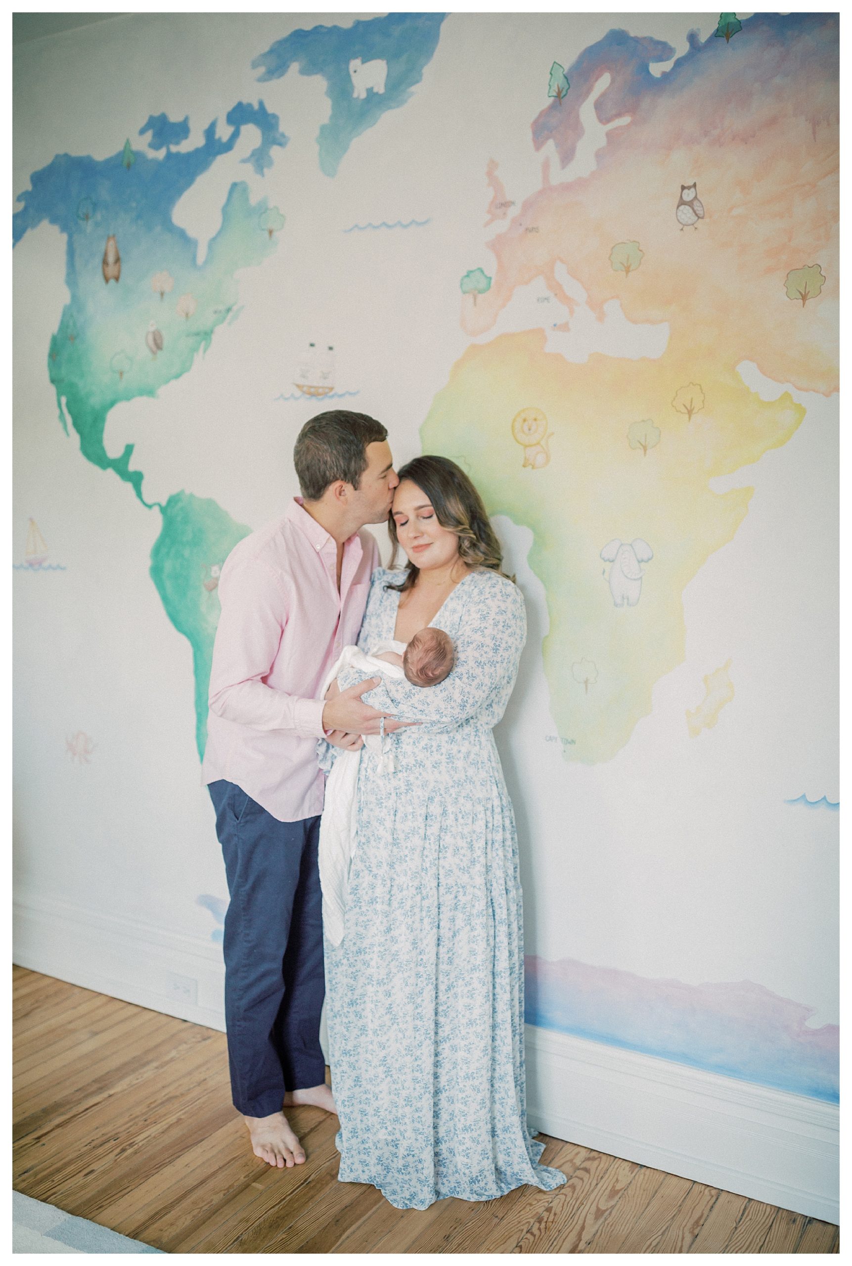 Father kisses mother's head while they stand in front of colorful mural in nursery, holding baby