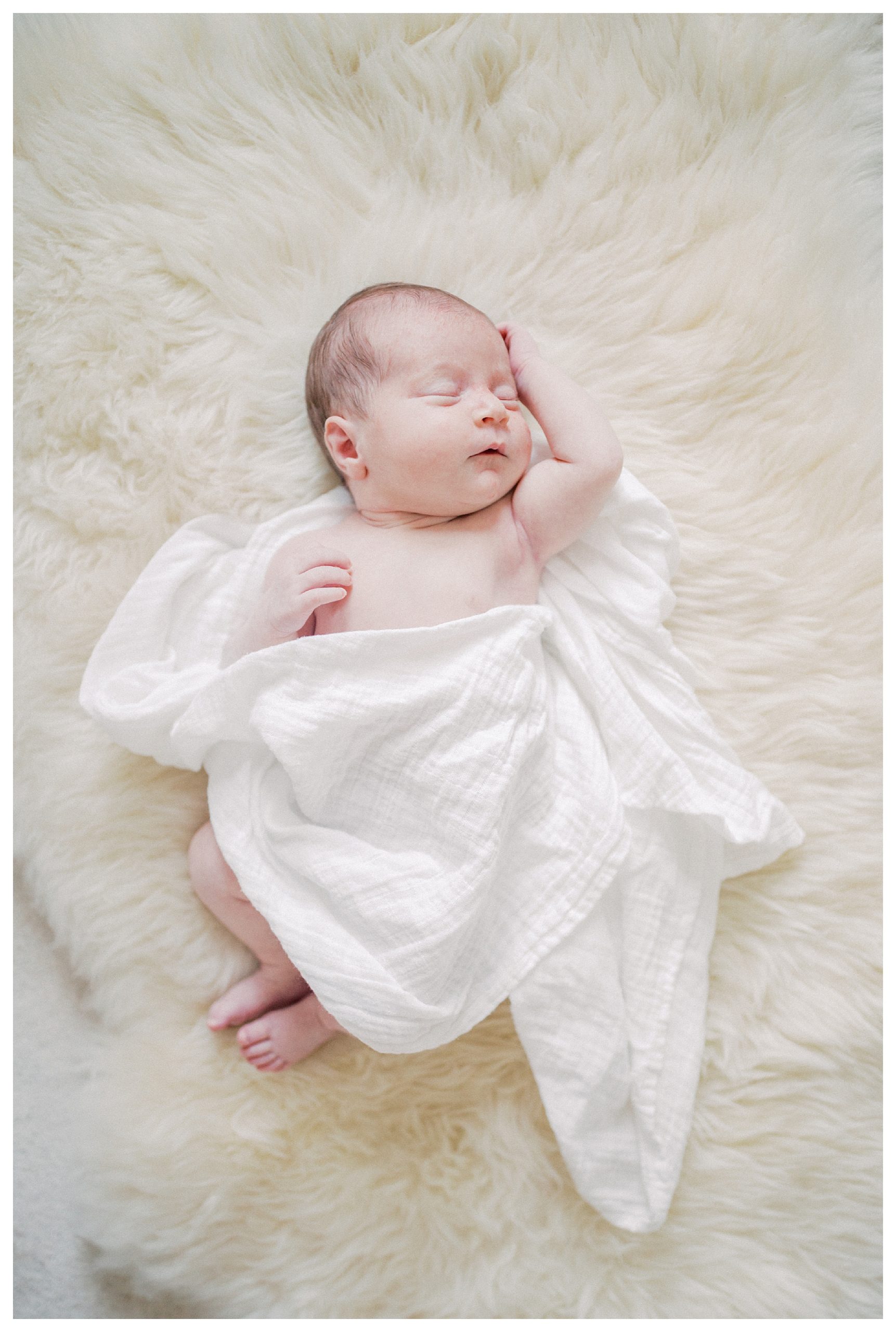 Newborn baby loosely swaddled in white peacefully sleeps on carpet during in-home newborn session.