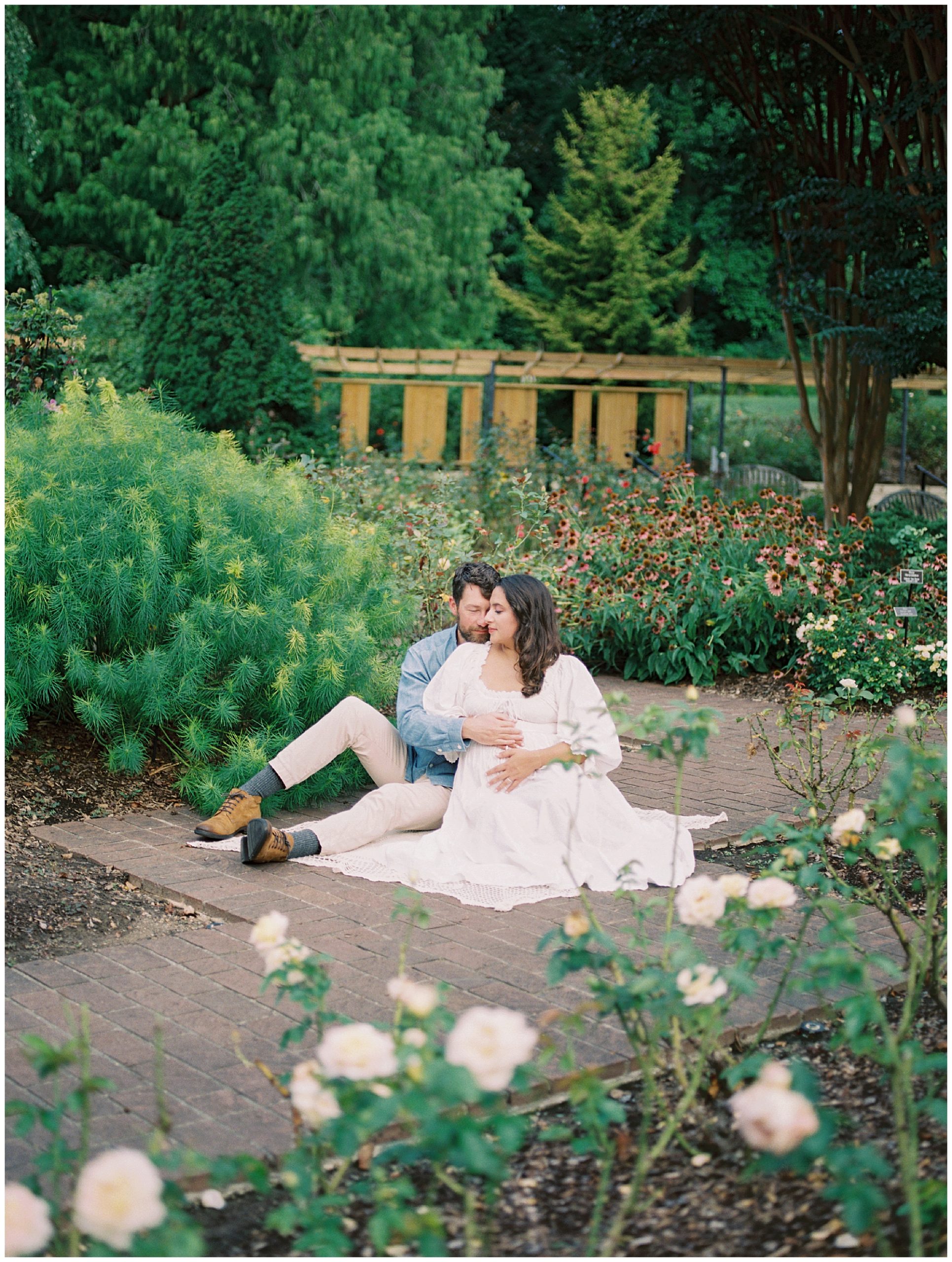 Husband and wife sit in the middle of a garden, embracing during their floral maternity session at Brookside Gardens.