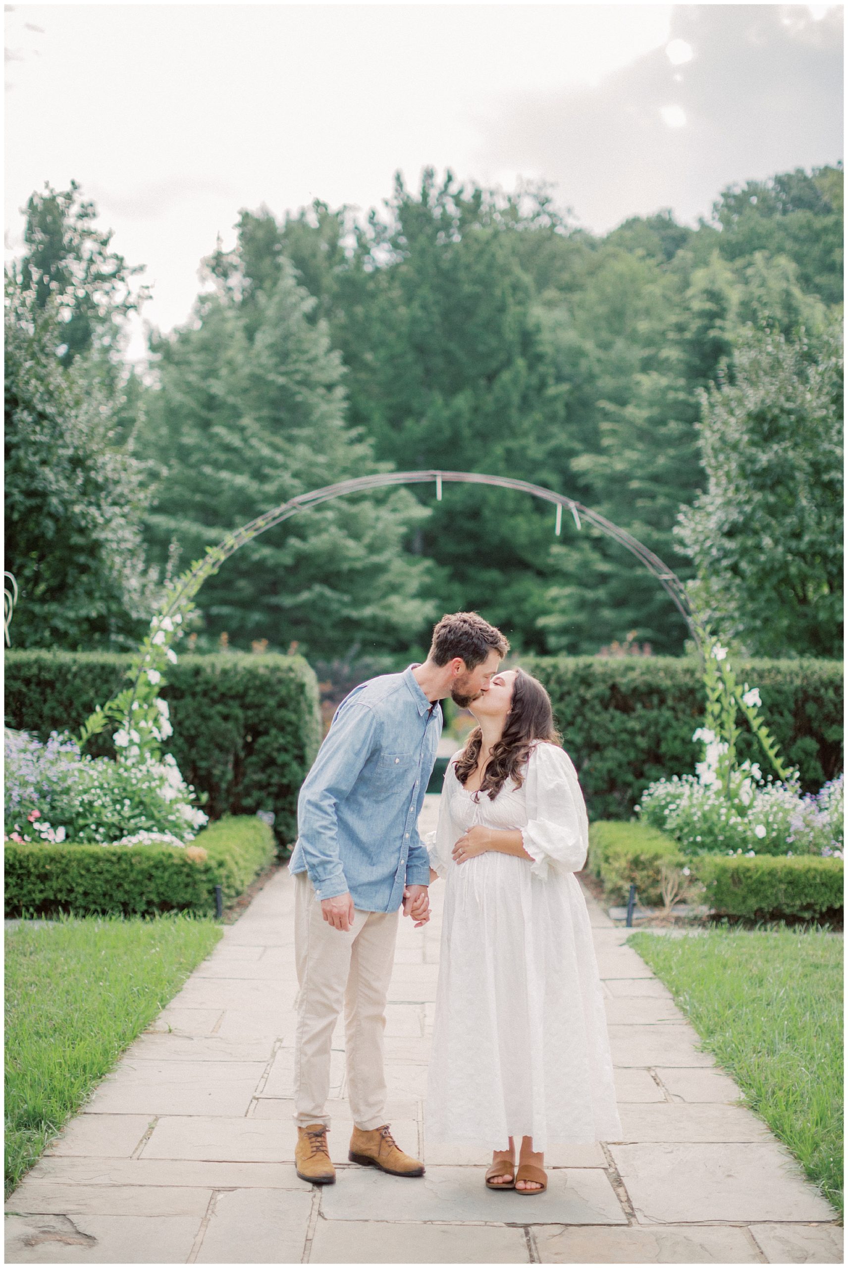 Couple stand together, holding hands and leaning in for a kiss, during their garden maternity session at Brookside Gardens.