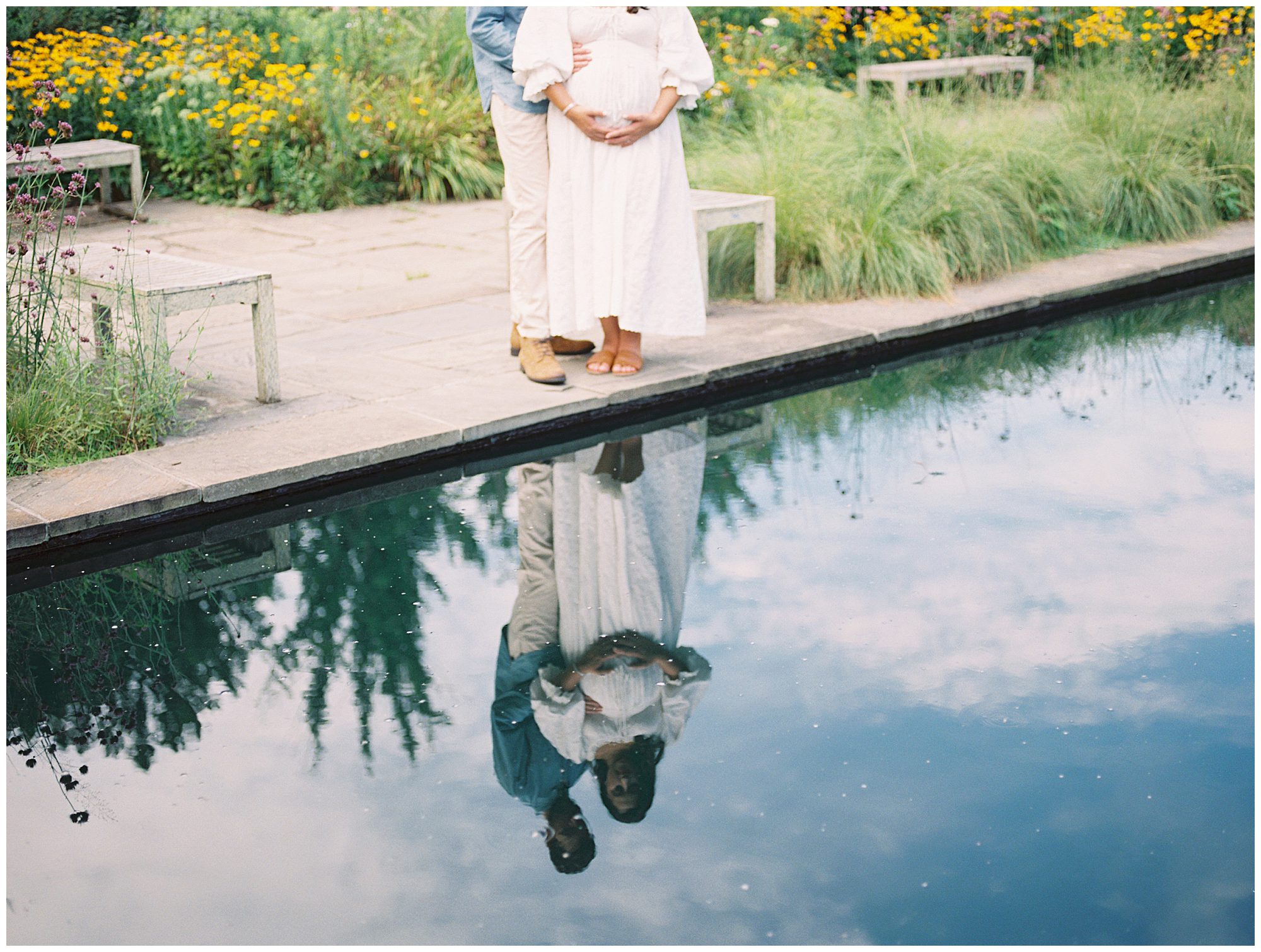 Reflection of expecting couple standing together at edge of a pond during their floral maternity session.