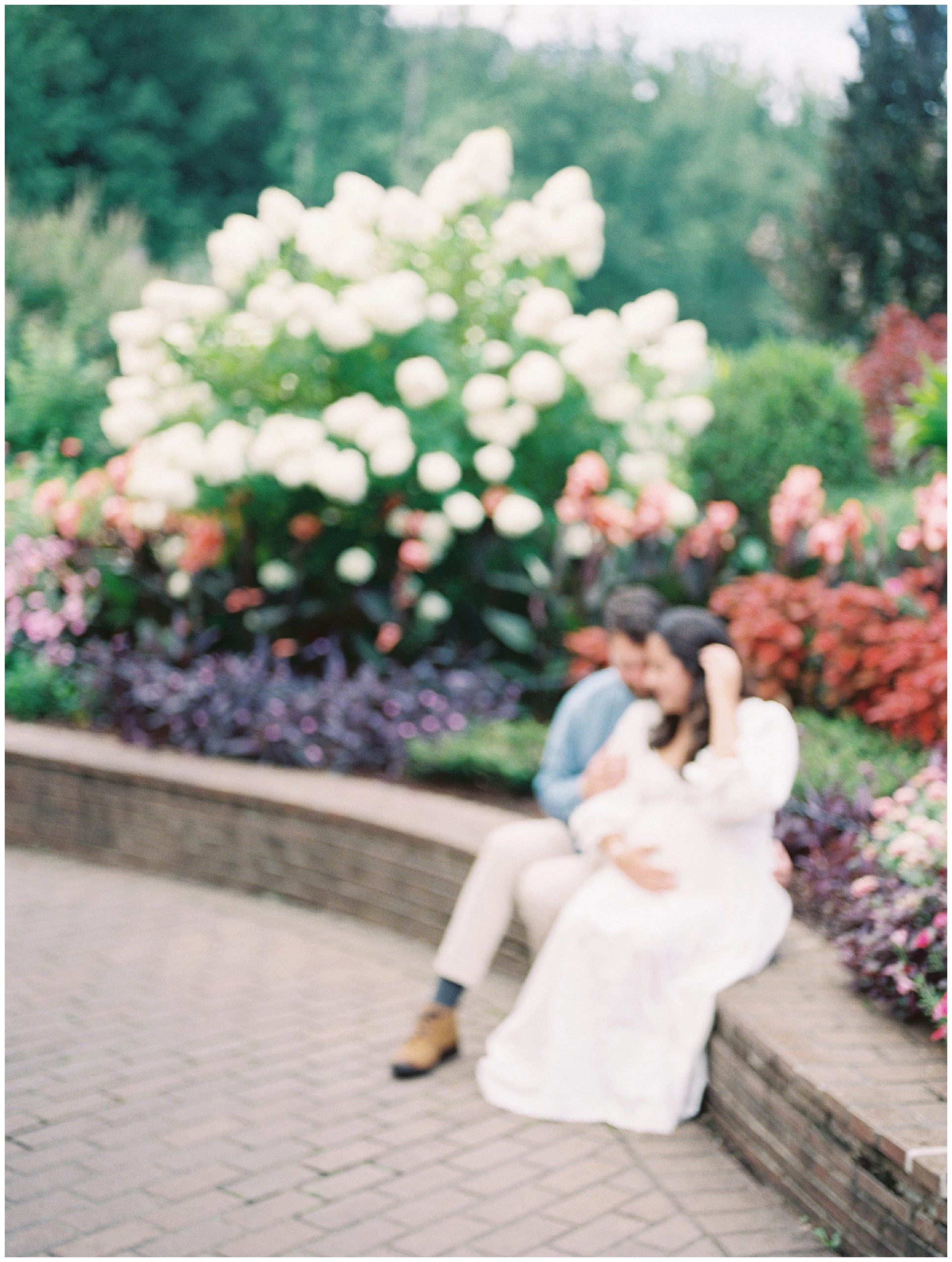 Out-of-focus image of couple sitting on brick wall in front of flowers at Brookside Gardens.