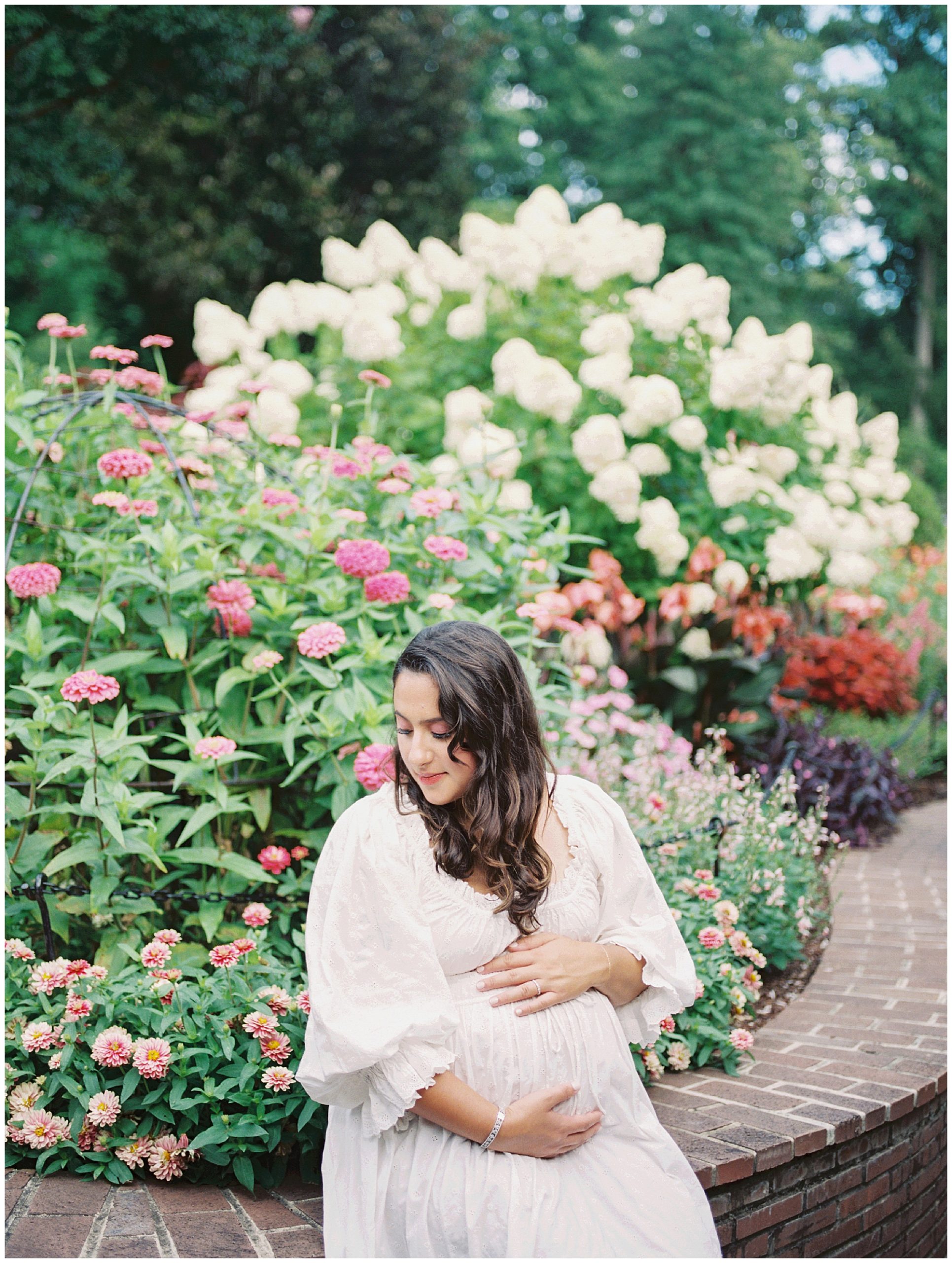 Pregnant mother sits on brick arch in front of pink and purple flowers during floral maternity session.