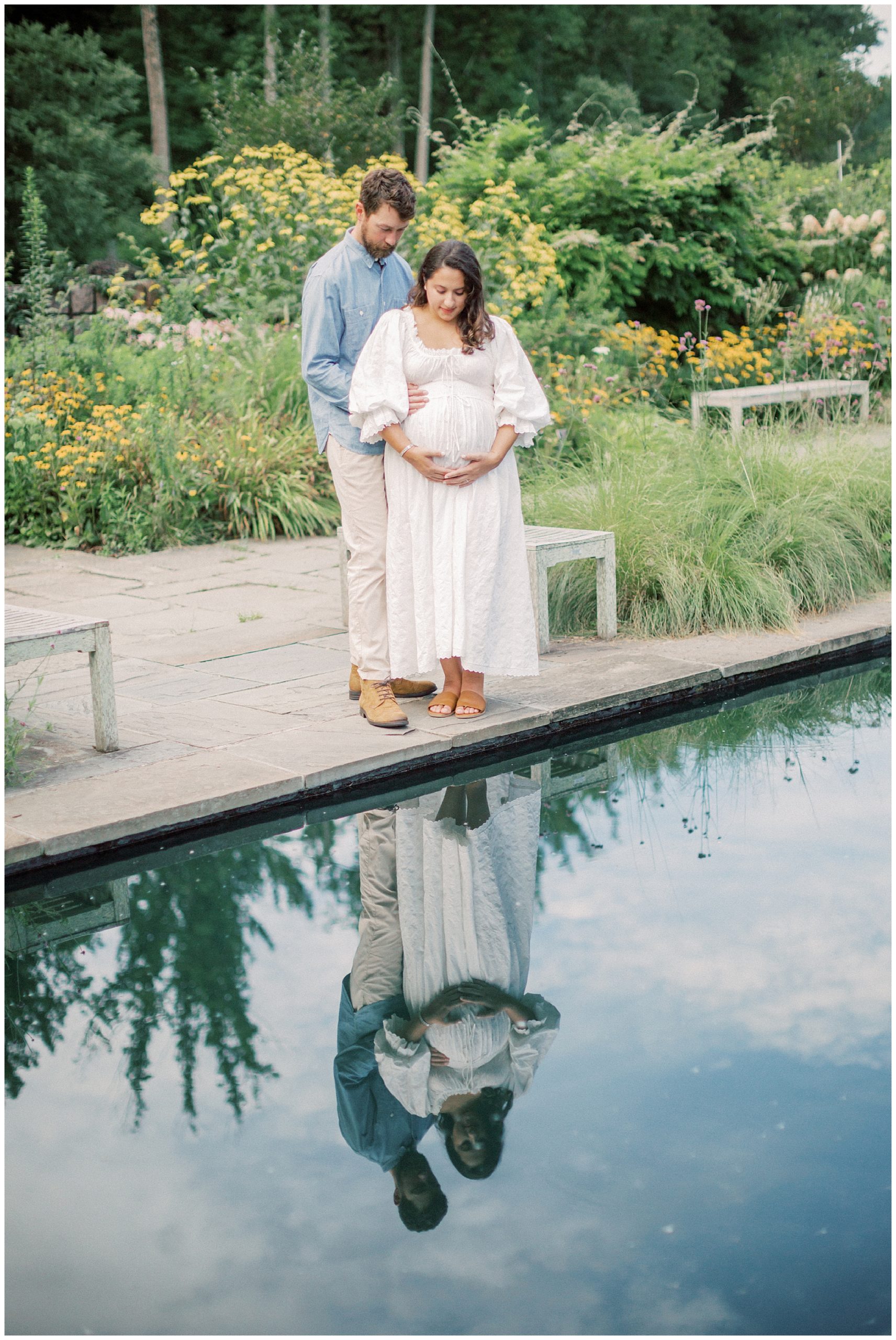 Couple stands at the edge of a pond in Brookside Gardens, looking down at mother's pregnant belly during their maternity session.