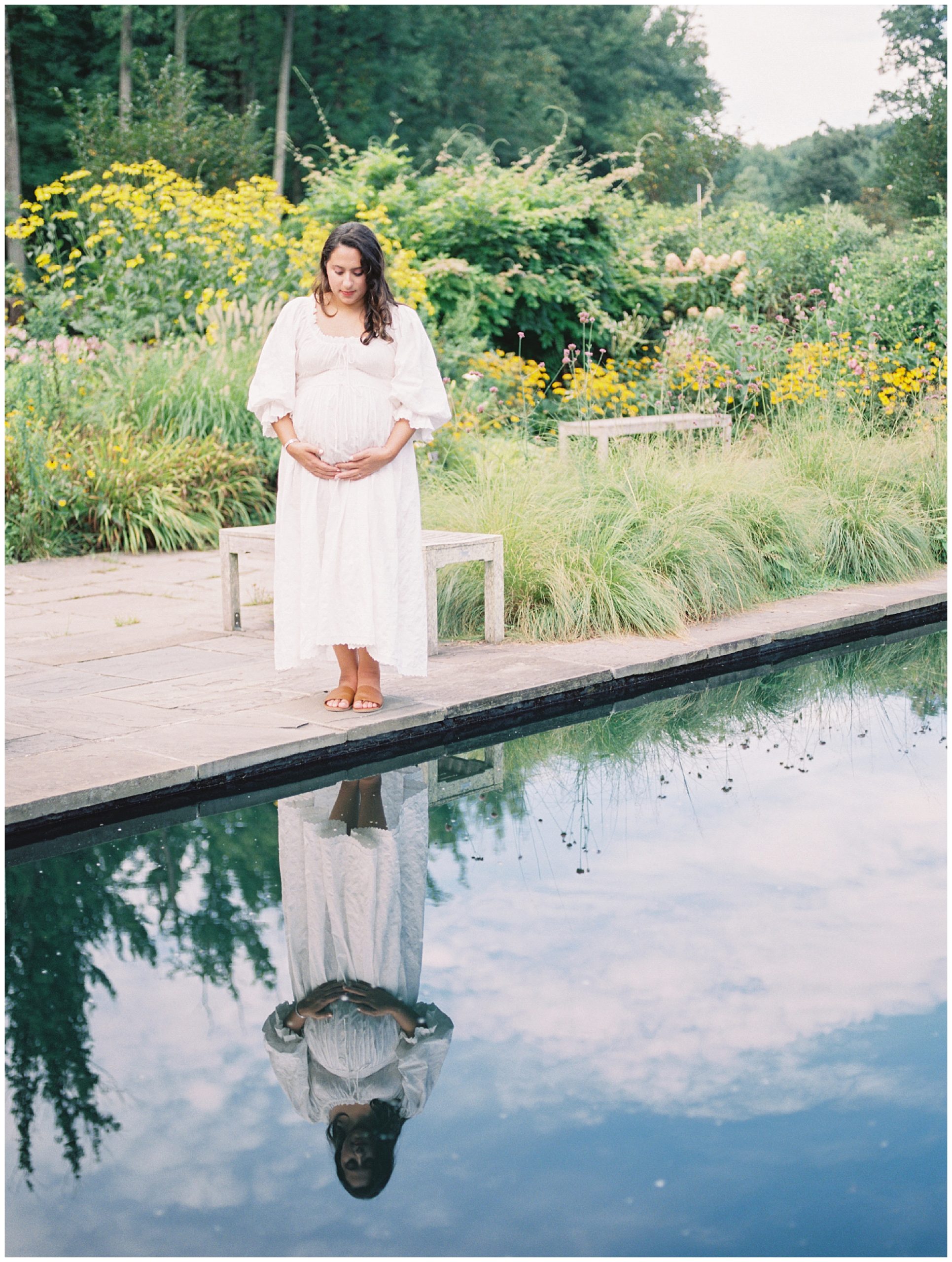 Pregnant mother in white Doen dress stands at the edge of a pond during her floral maternity session.