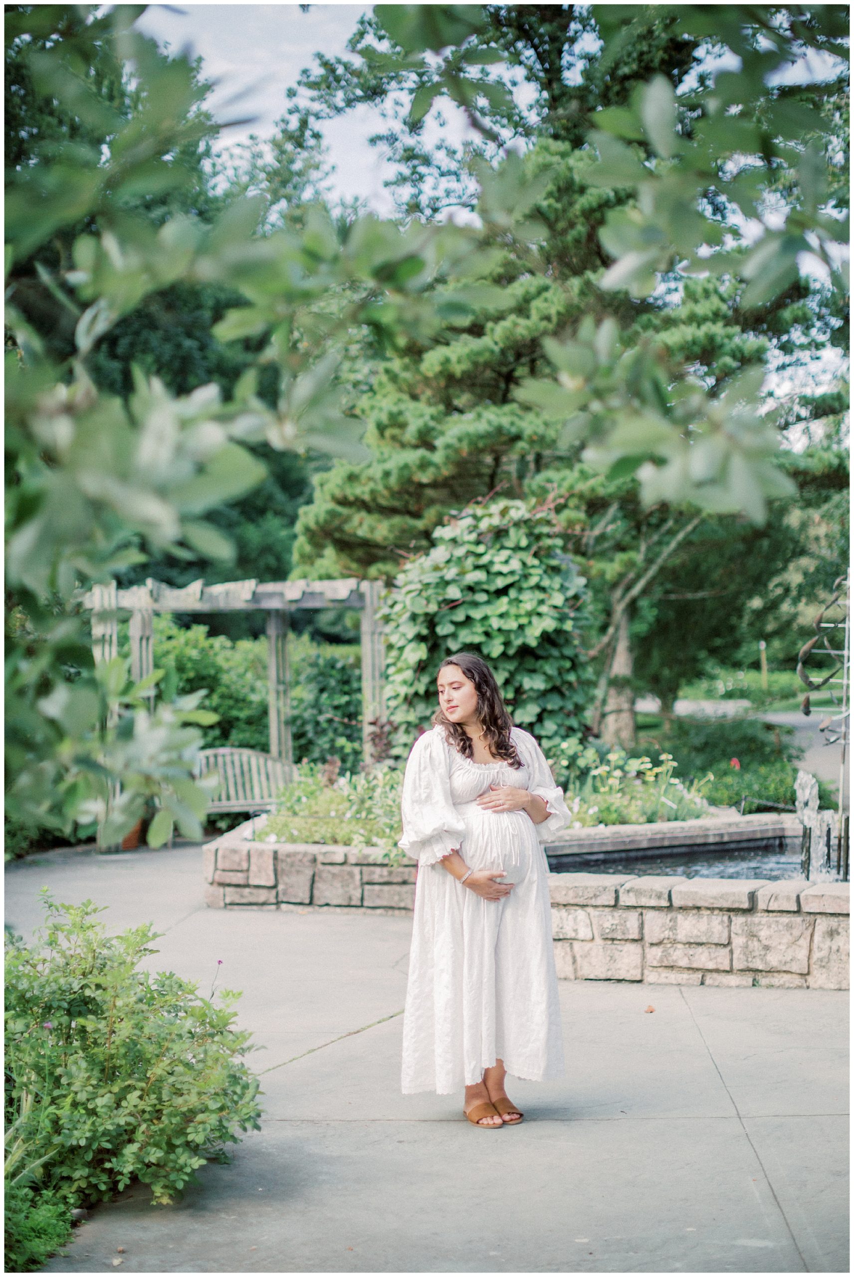 Pregnant mother in white Doen dress places one hand on top and one hand below her belly while standing in a green garden.