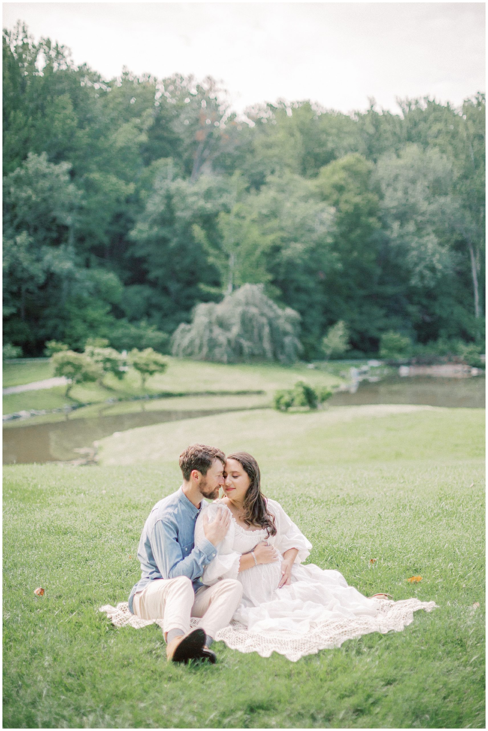 Expecting father and mother sit on blanket in an open lawn during their maternity session.
