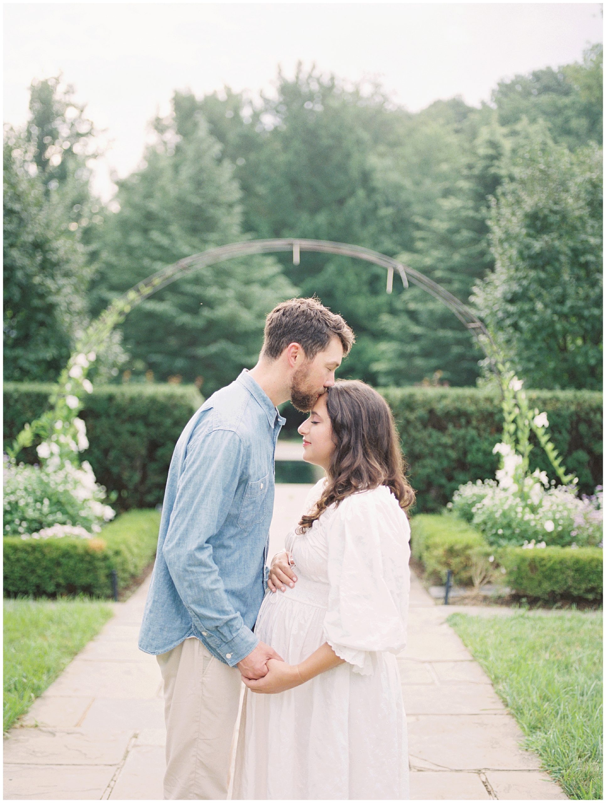 Expecting father kisses the top of his wife's head as they stand by a floral arch during their garden maternity session.