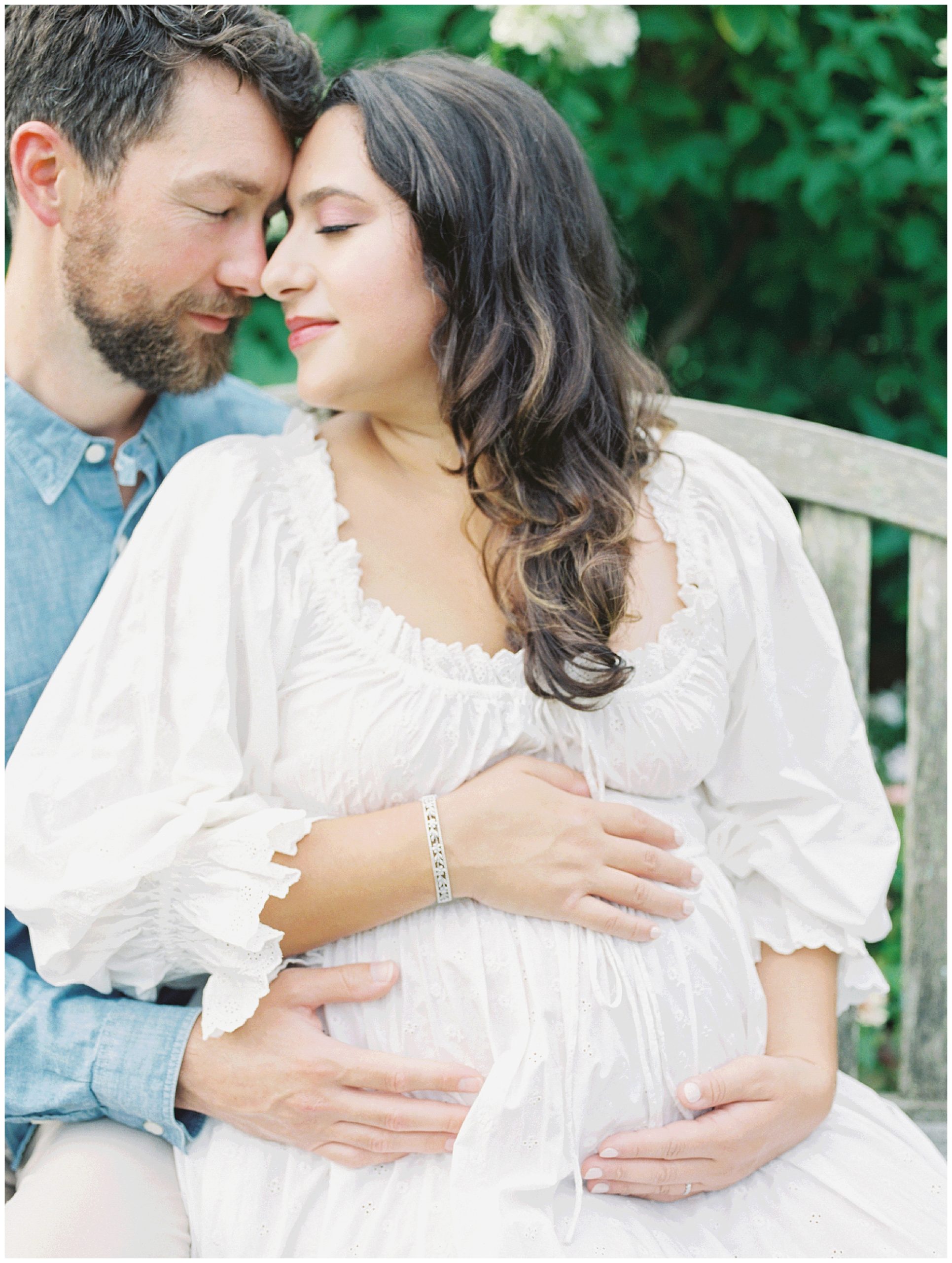 Expecting parents sit together on a bench, leaning in to one another, during their maternity session at Brookside Gardens.