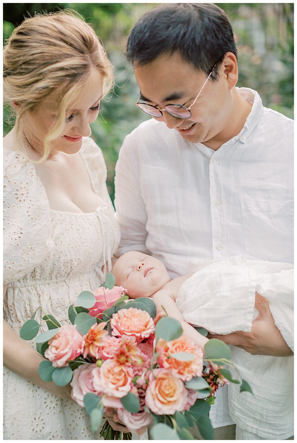Newborn baby sleeps on bed of roses while being held by parents during outdoor newborn session.