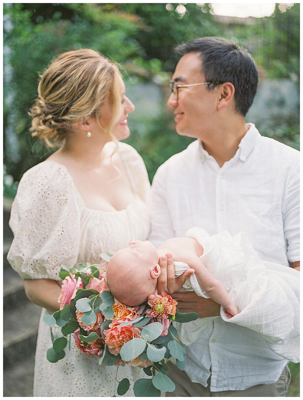 New parents hold their newborn daughter with bouquet of roses and smile at each other.