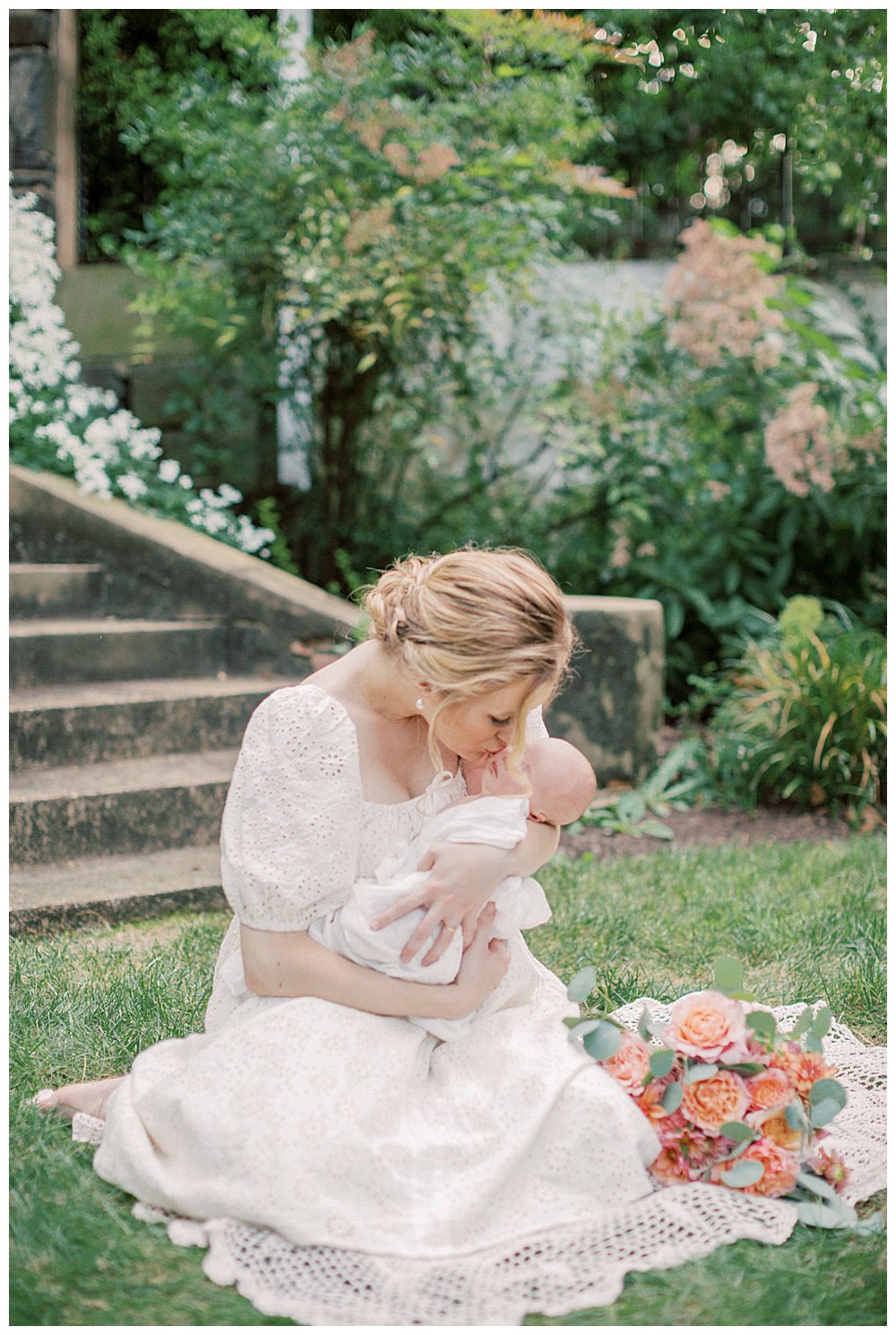 Blonde new mother holds newborn daughter while sitting on crochet blanket during outdoor Alexandria VA newborn session.