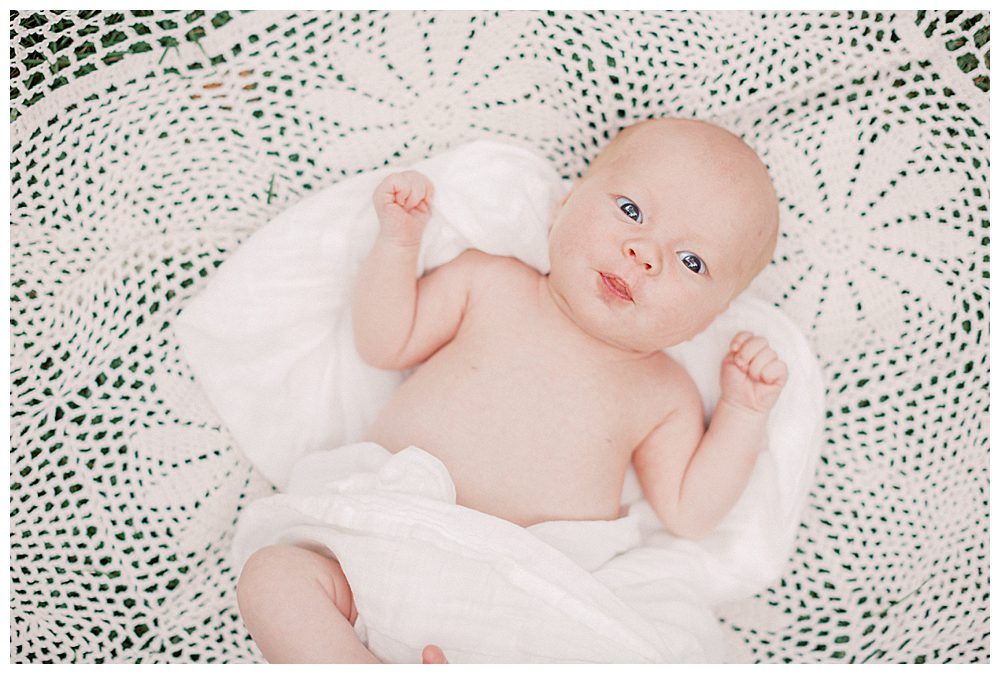 Newborn baby looks up at camera while laying on crochet white blanket outside.