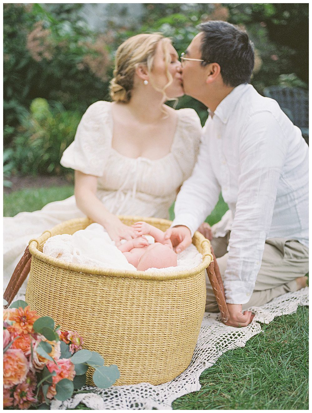 New parents lean in for a kiss while placing their hands on newborn daughter in a Moses basket during outdoor newborn session.