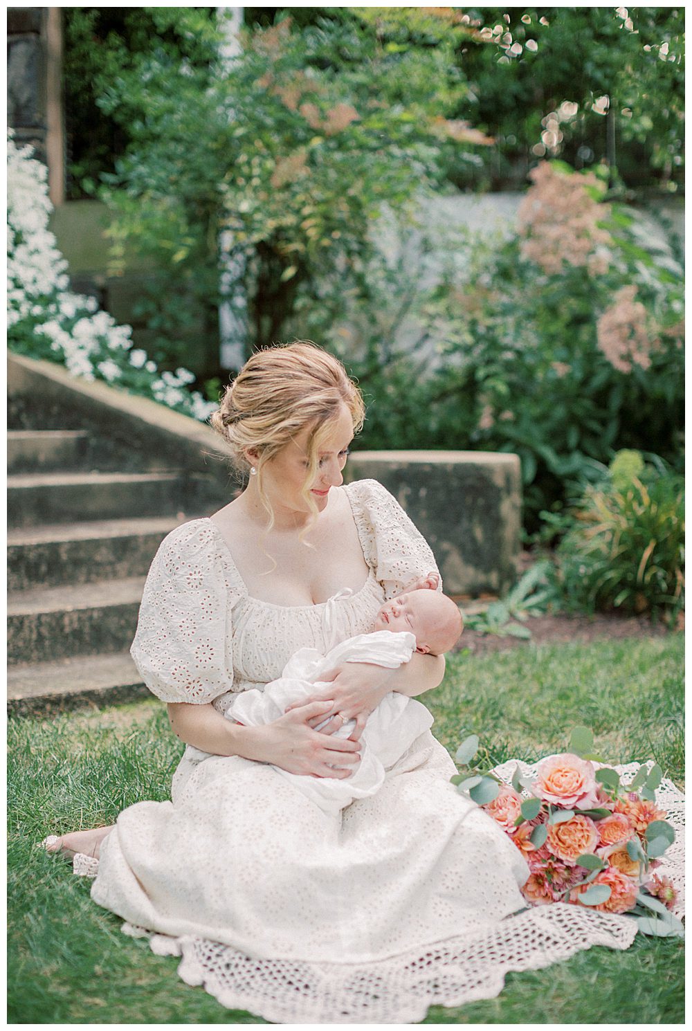 Mother holds newborn daughter while sitting on blanket with roses outside during Alexandria VA newborn session.