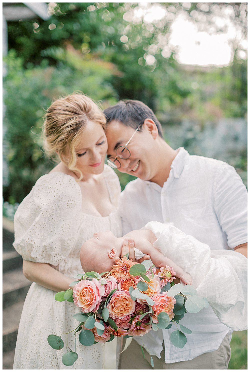 Father holds newborn daughter while leaning into his wife who holds a bouquet of roses during outdoor newborn session.