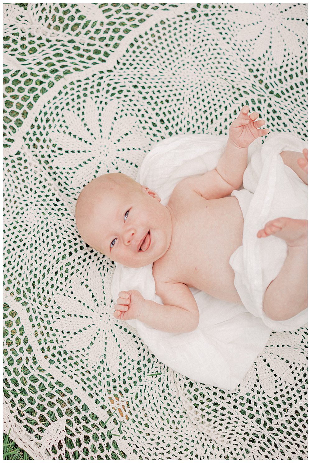 Newborn baby girl smiles as she lays on crochet blanket outside.