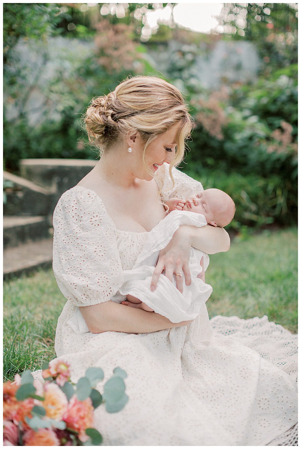 Blonde mother holds and smiles down at newborn daughter as she sits on blanket outside.