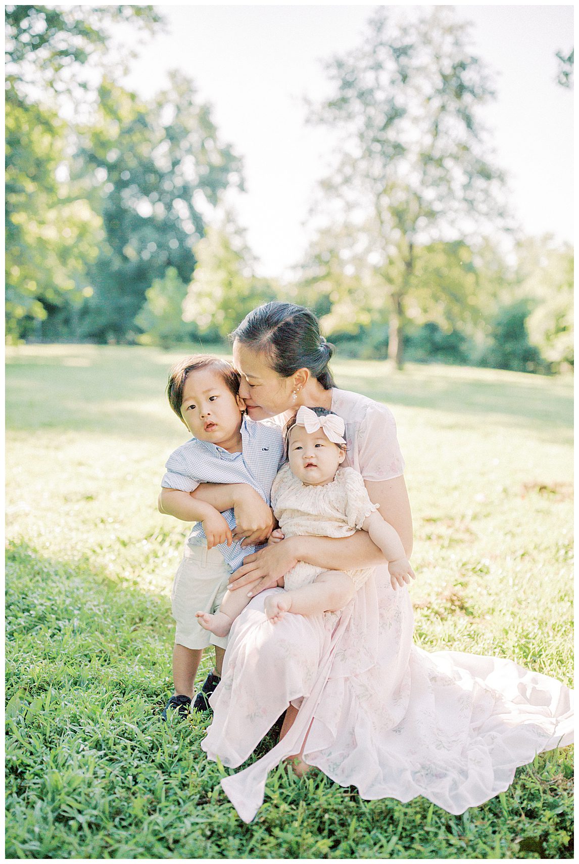 Mother in pink dress kneels down and kisses her toddler son's cheek while her infant daughter sits on her lap.