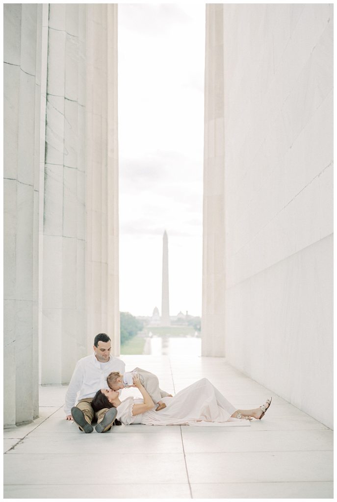 Woman lays in husband's lap while kissing baby boy in the Lincoln Memorial.