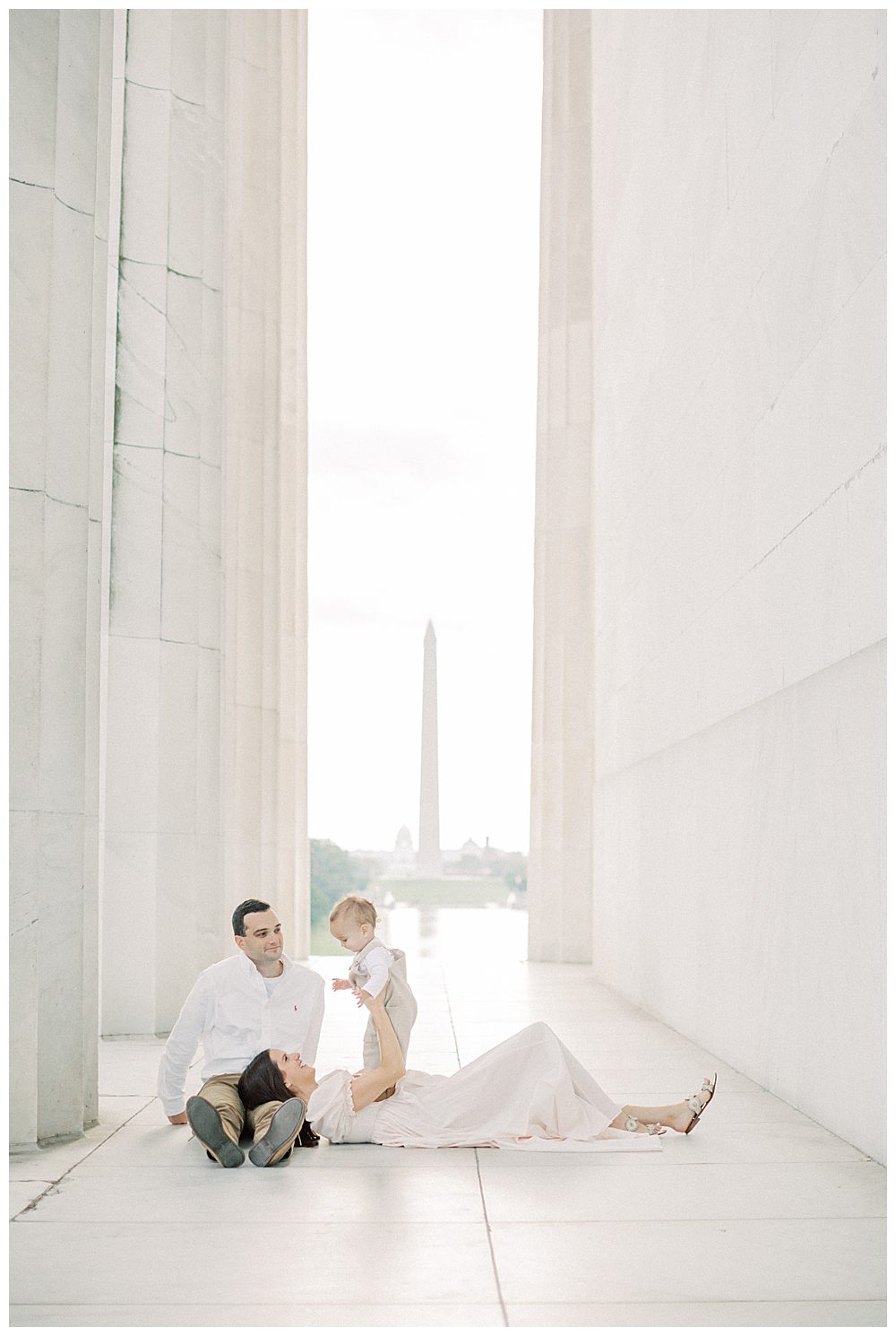 Mother holds up toddler son while laying in husband's lap at Lincoln Memorial during DC Monuments Family Photo Session.