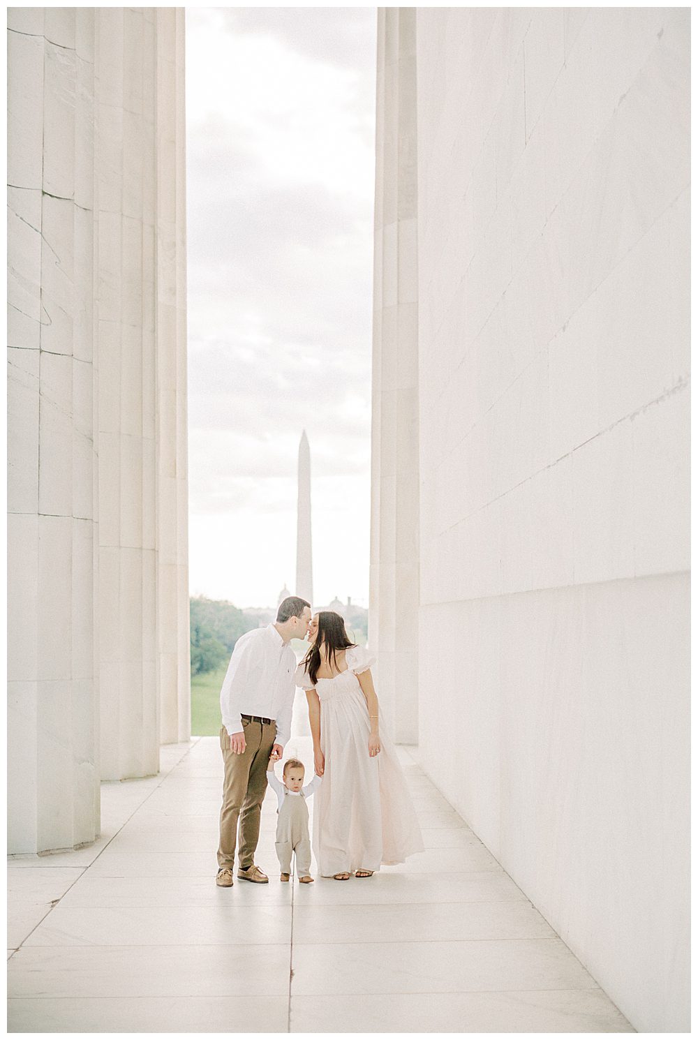 Parents hold their toddler's hands and lean in to kiss while at the Lincoln Memorial.