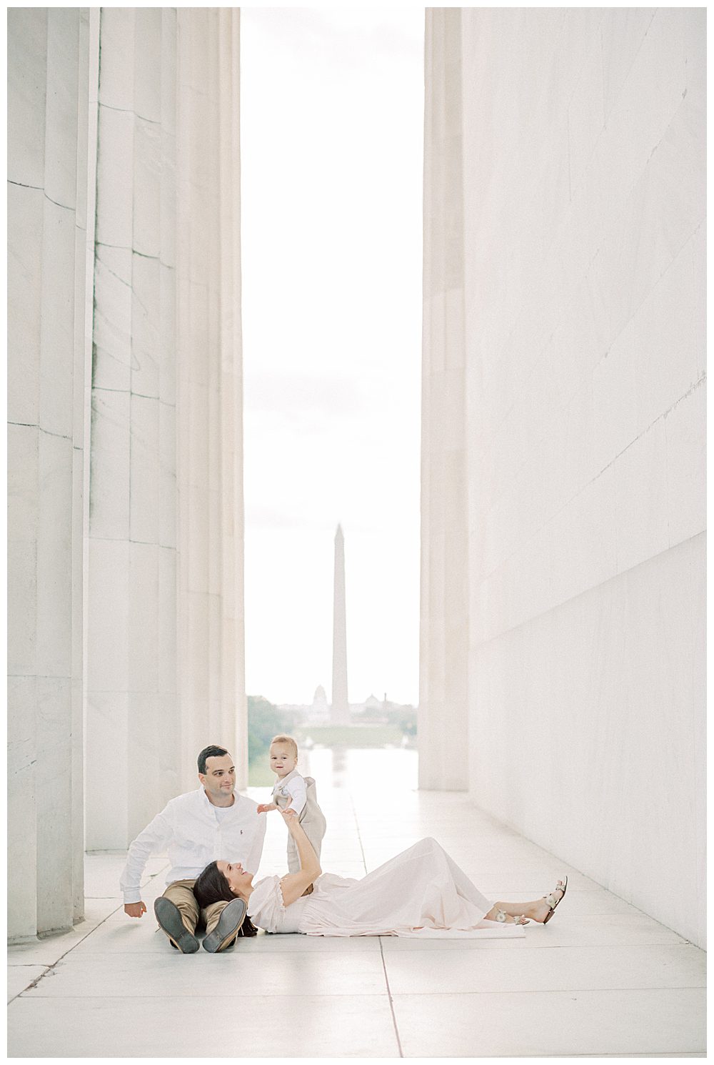 Mother lays in her husband's lap as she holds up her toddler son at the Lincoln Memorial during DC Monuments Family Photo Session.